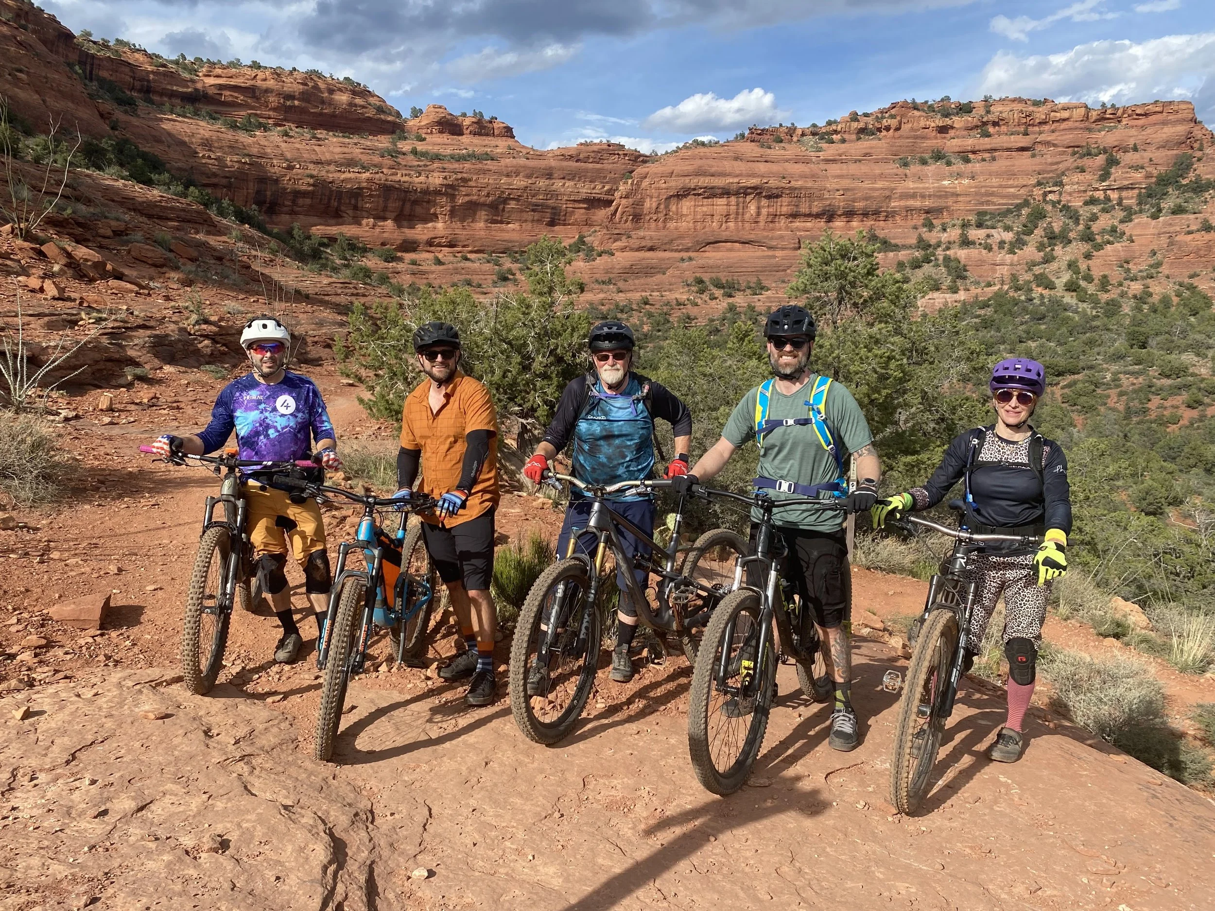 Five mountain bikers in cycling gear, wearing helmets and sunglasses, standing with their bikes on a dirt trail in a desert landscape with red rock formations and sparse vegetation.