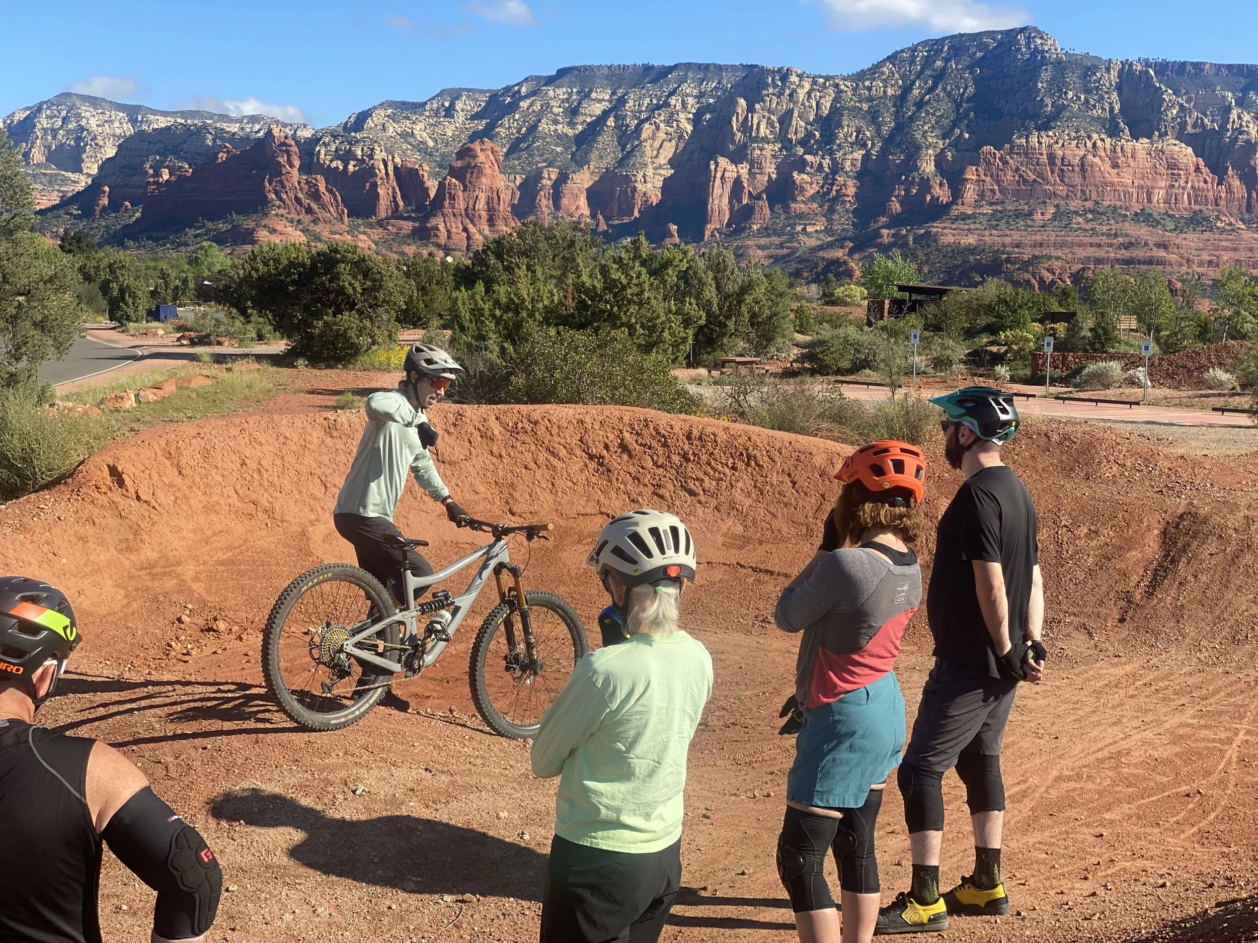 Group of mountain bikers at a dirt bike park pump track with red rock formations and mountains in the background, learning from a certified mountain bike instructor.