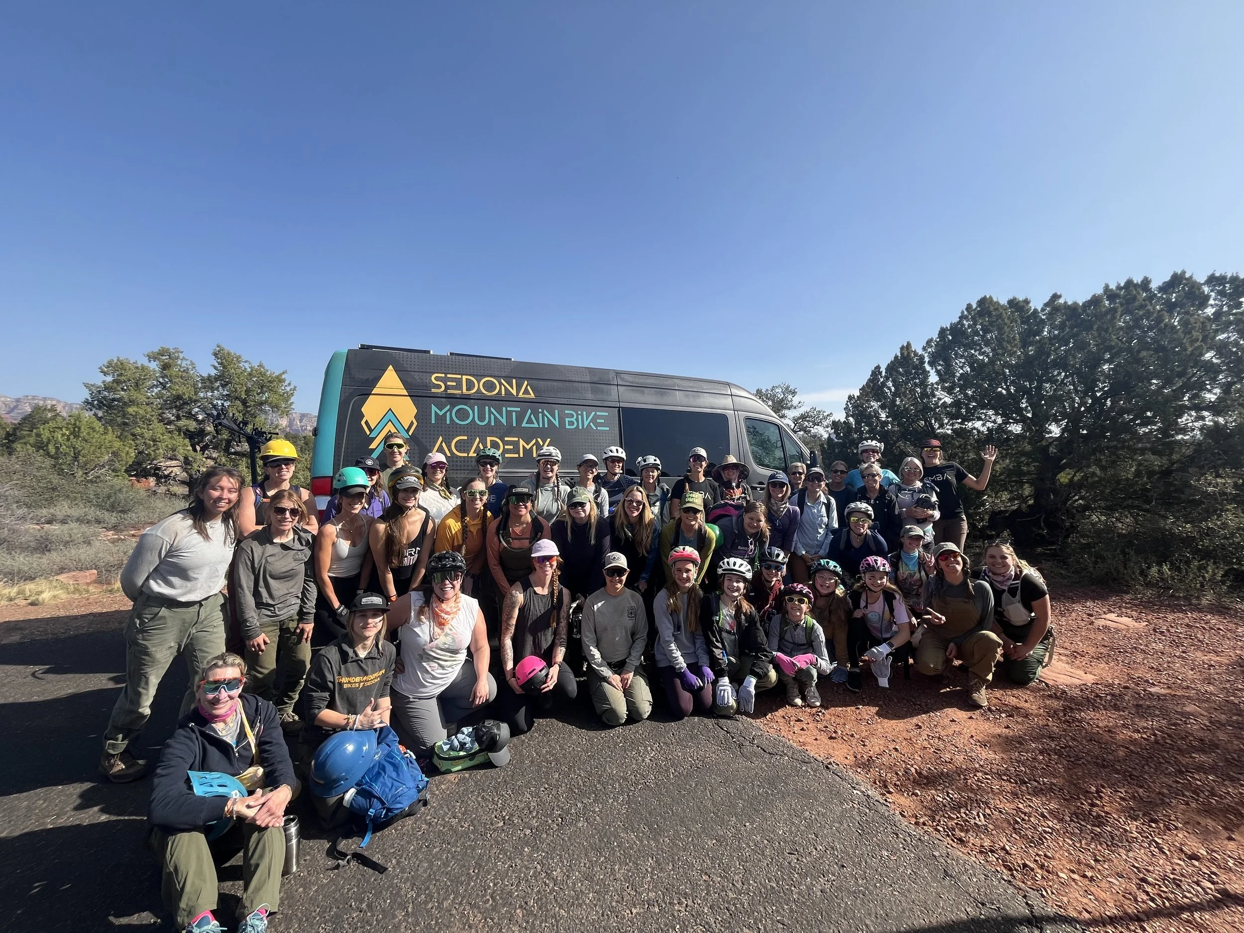 Group of women wearing helmets and outdoor clothing, posing in front of a van with 'Sedona Mountain Bike Academy' logo, with trees and red rock terrain in the background, preparing to conduct volunteer trailwork.