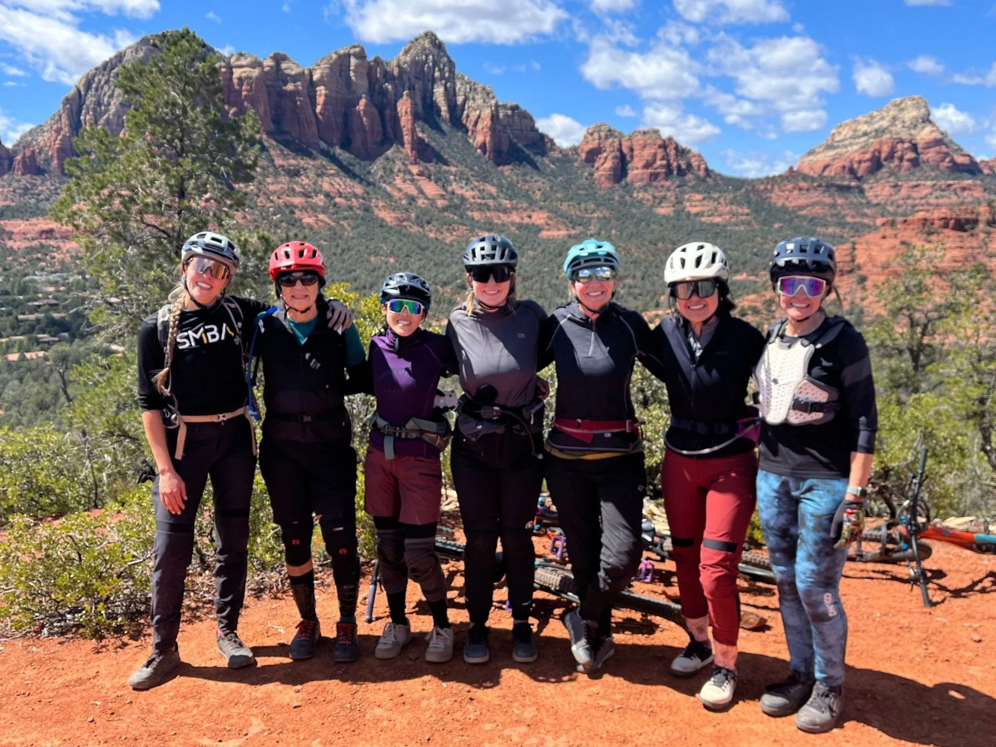 Seven women in cycling gear and helmets standing together on a dirt trail with desert landscape, red rocks, and mountains in the background on a sunny day, enjoying the benefit of quality instruction and a local guide.