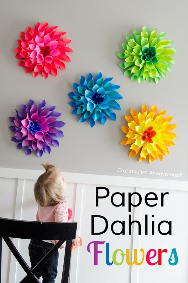 Large dahlias made from colorful papers decorate a wall. A toddler looks up at them.