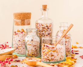 A collection of glass containers filled with rose petal bath salts sits amongst wooden spoons and dried flower petals