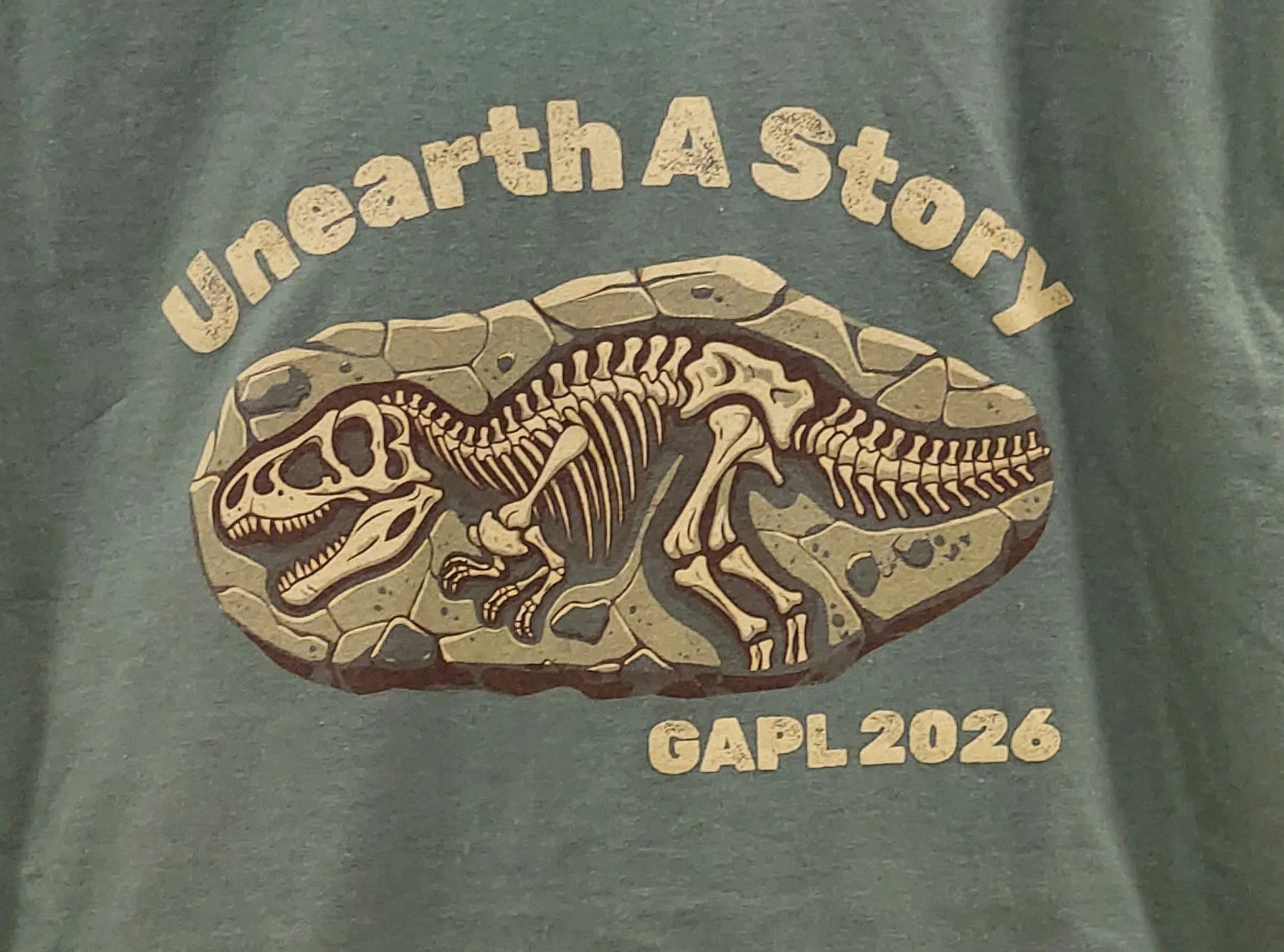 T-shirt detail Unearth a Story GAPL 2026. Words around a fossil of a complete T-Rex skeleton in stone (Copy)