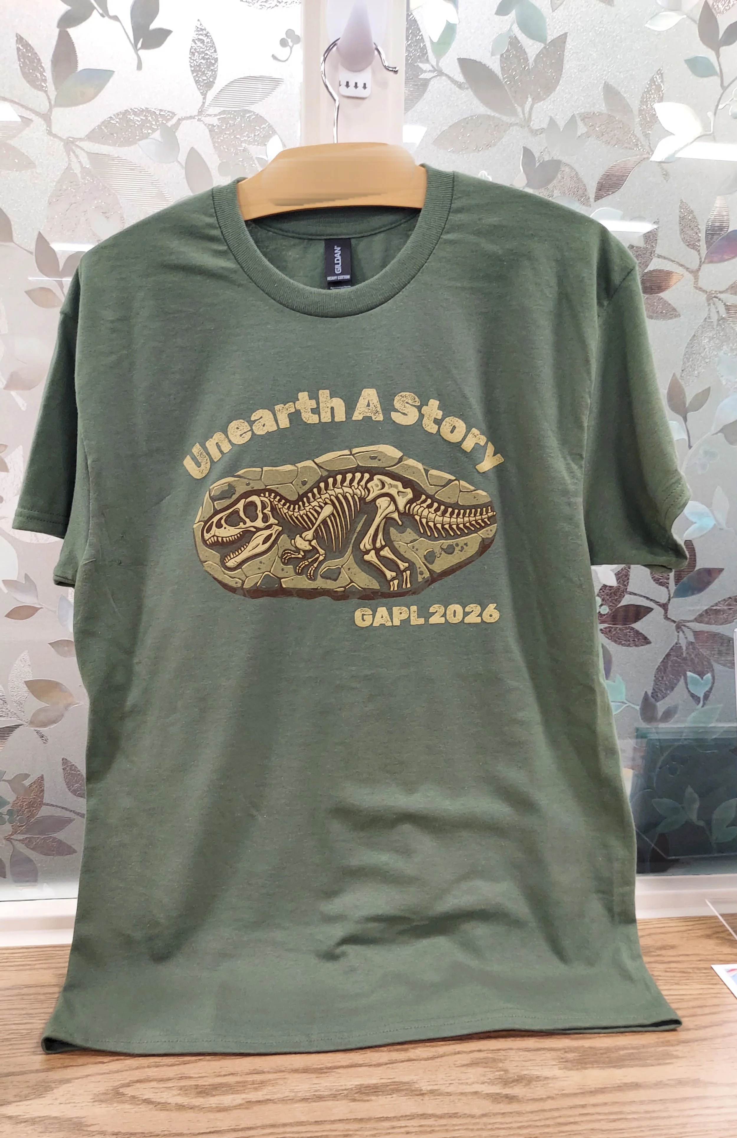 Summer Reading Tshirt Unearth a Story GAPL 2026. Words around a fossil of a complete T-Rex skeleton in stone (Copy)