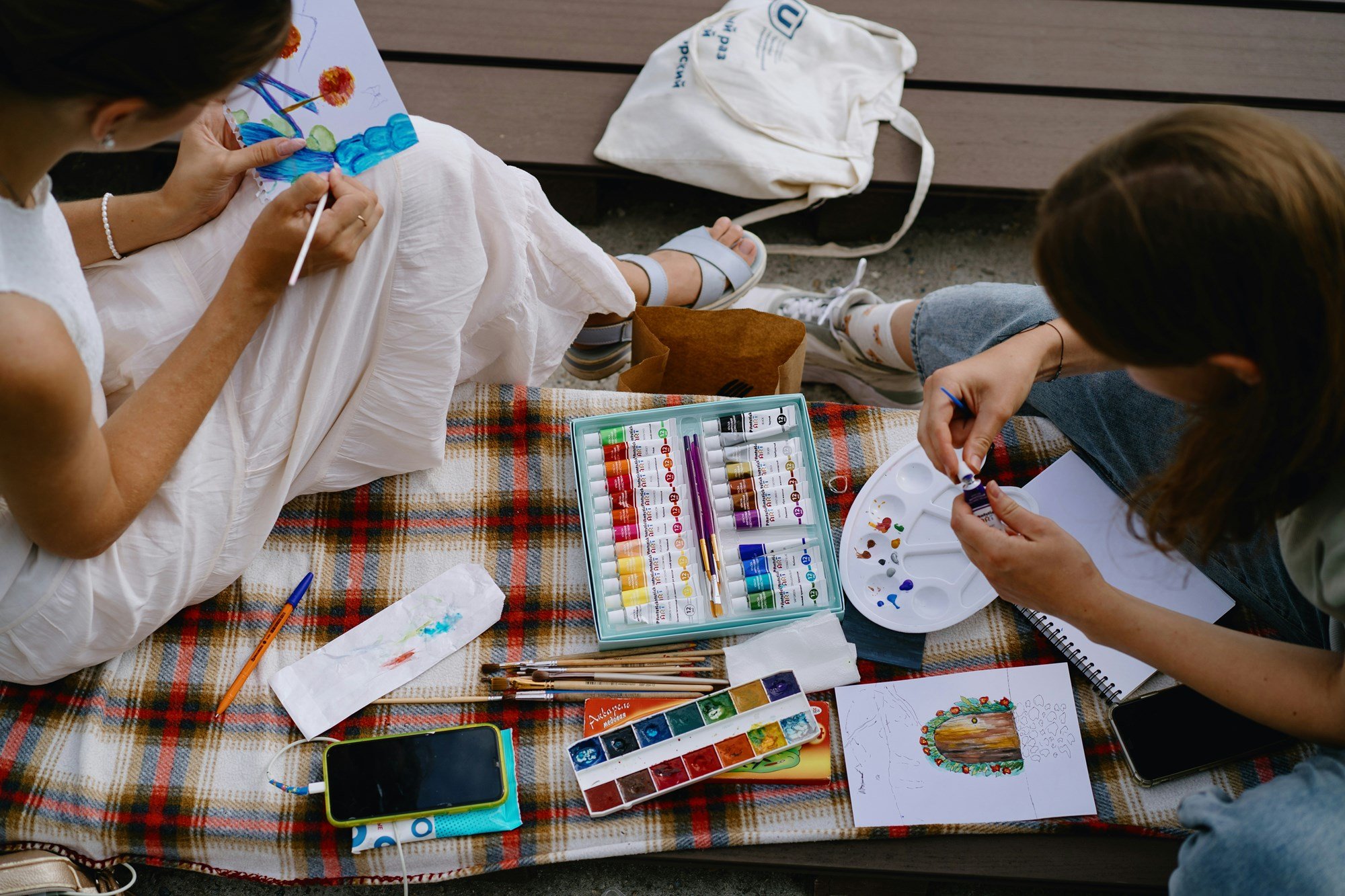Two young women share water color and art supplies between them on a plaid blanket
