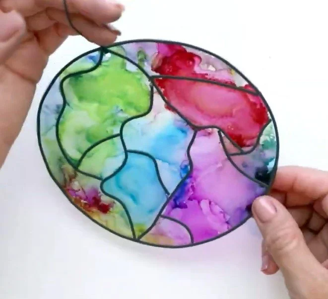 A circular, colorful suncatcher made from acetate and alcohol ink with adhesive leading is held between two hands.