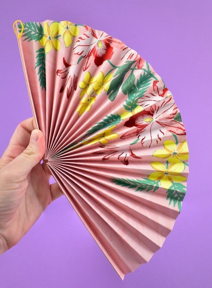 A white hand holds a pink, floral paper fan open on a purple background