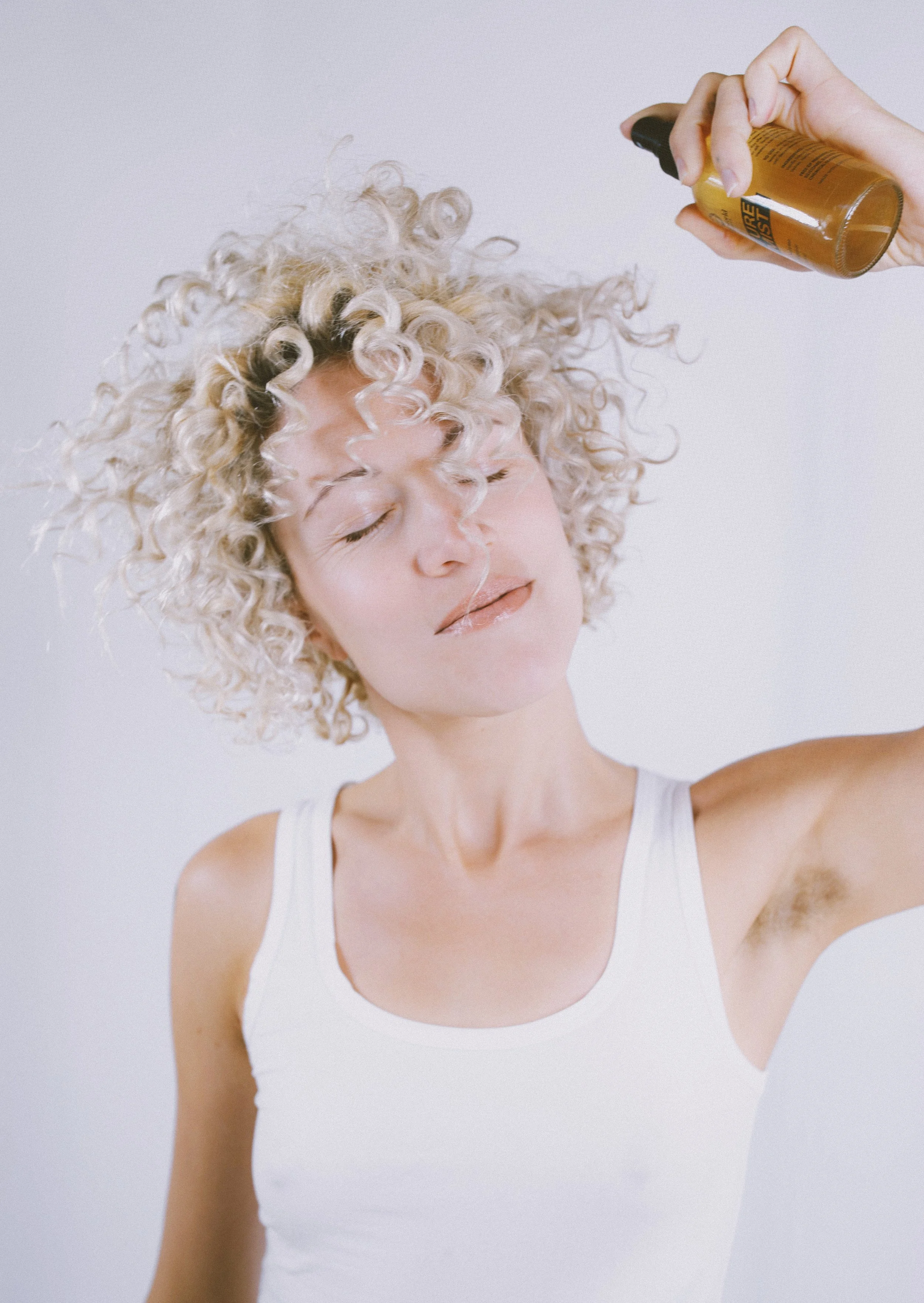 Woman with curly blonde hair eyes closed, wearing a white tank top, applying a spray product to her hair.