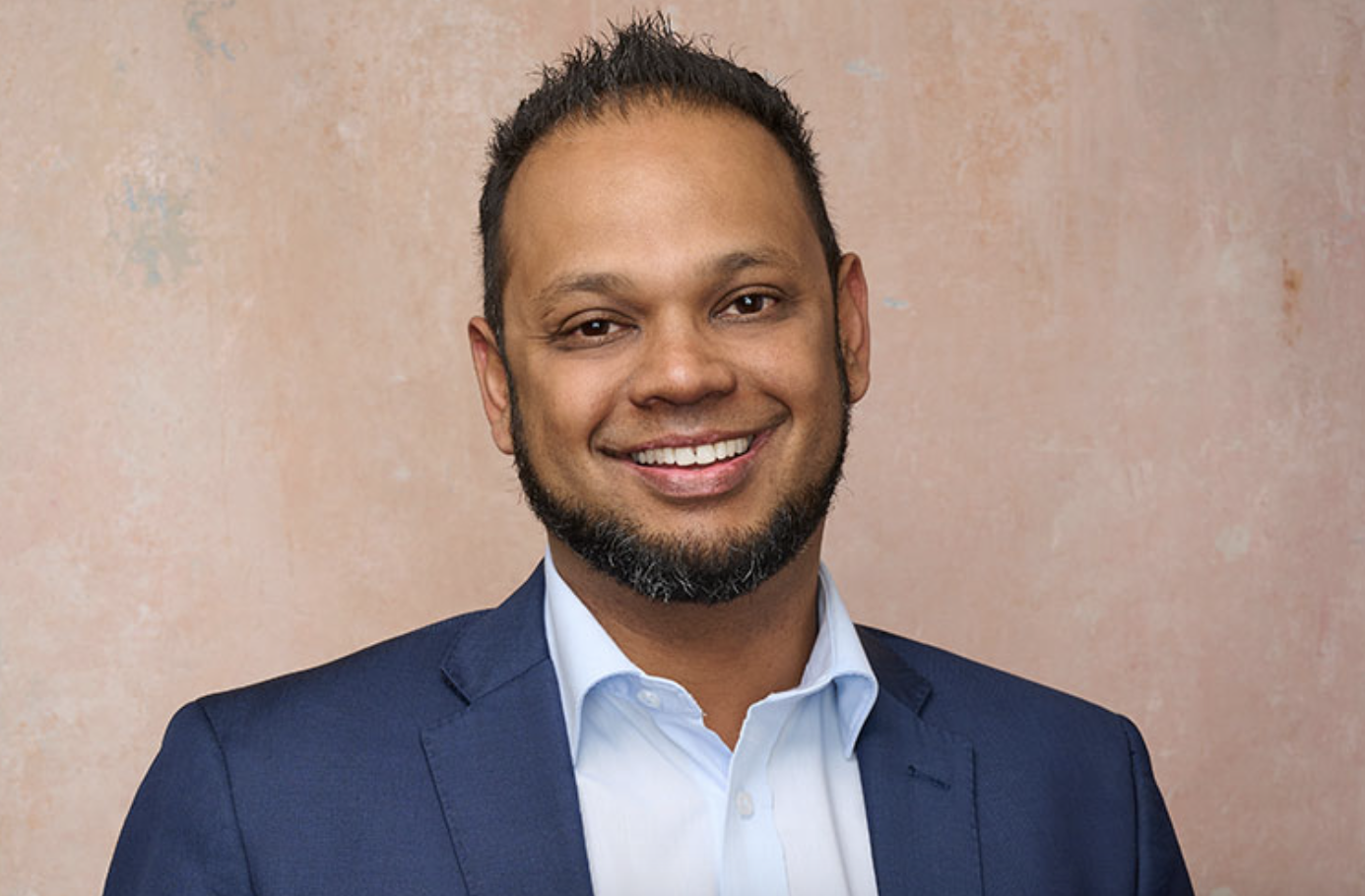 A smiling man with a beard and short hair, wearing a navy blazer and light blue shirt, standing against a textured beige background.