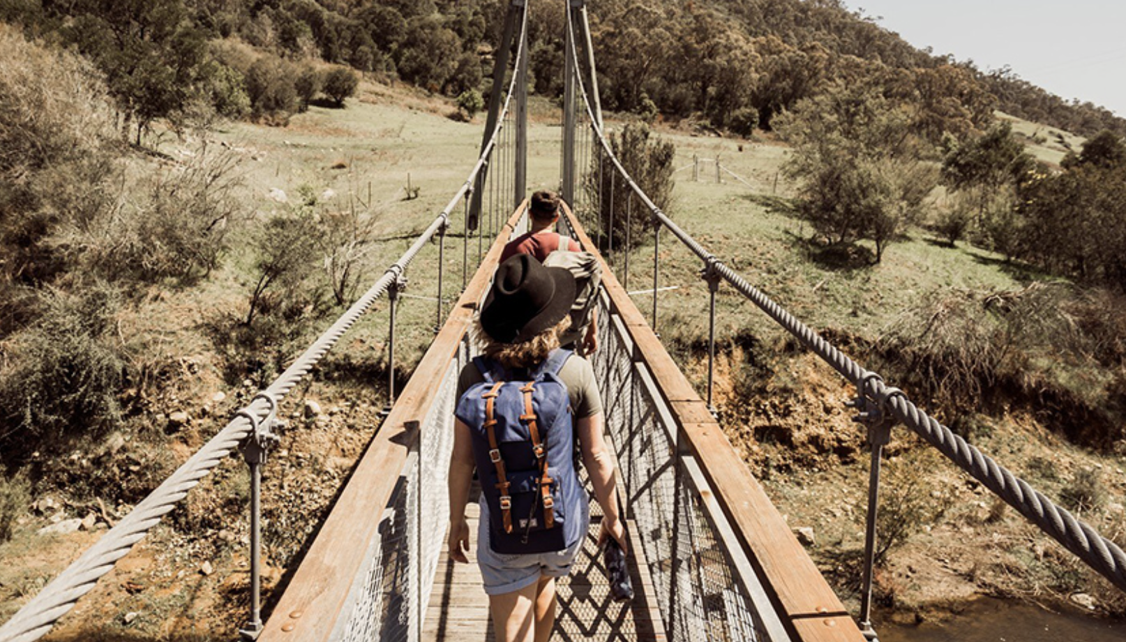 Three hikers walking across a suspension bridge in a hilly, wooded area during daytime.