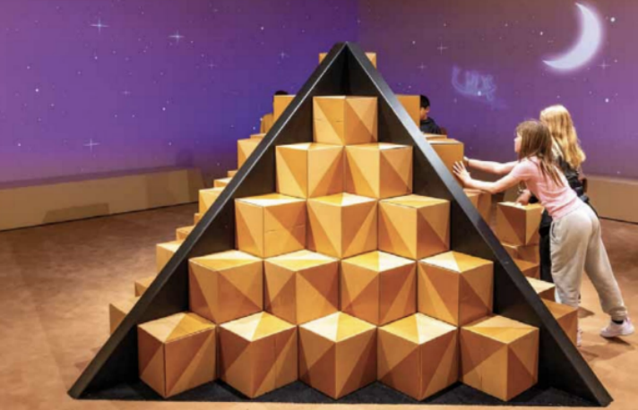 Children playing with large orange pyramid-shaped blocks arranged in a pyramid inside a room with a purple night sky and moon mural.