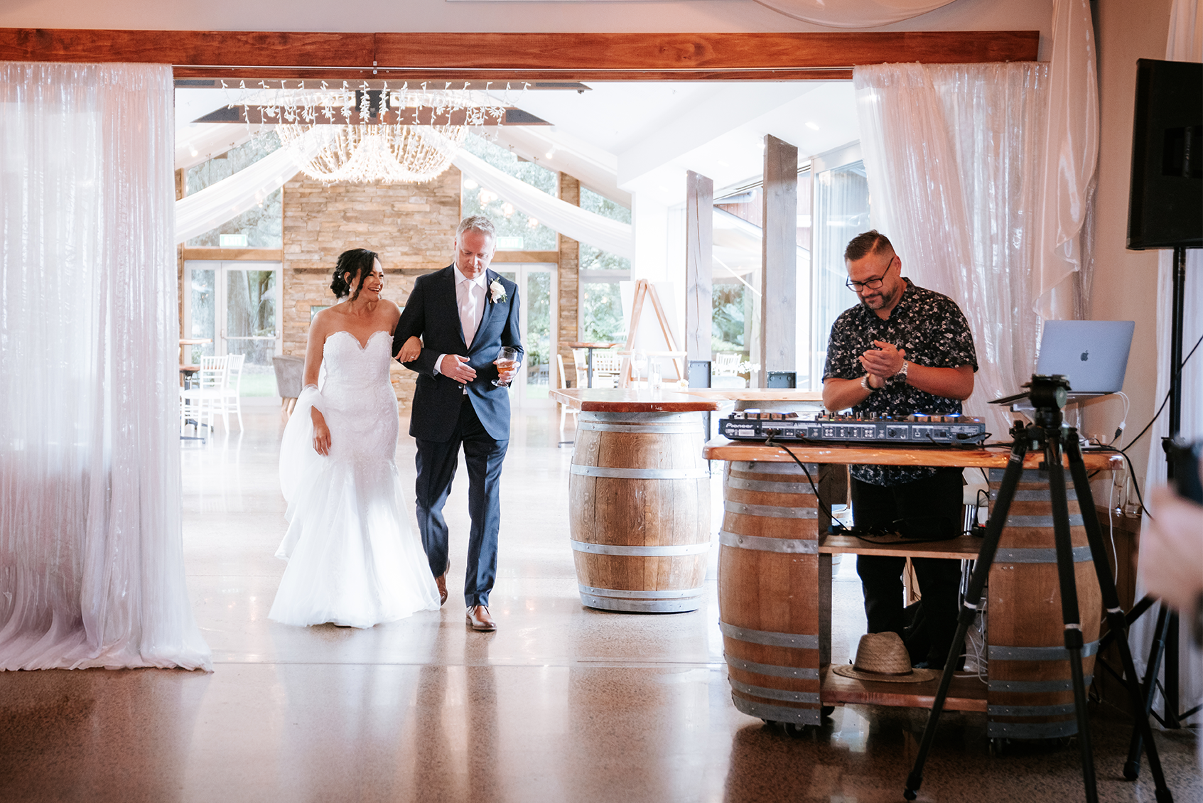 A bride and groom walking arm in arm at their wedding reception, with a DJ and audio equipment nearby in a decorated venue with natural light.