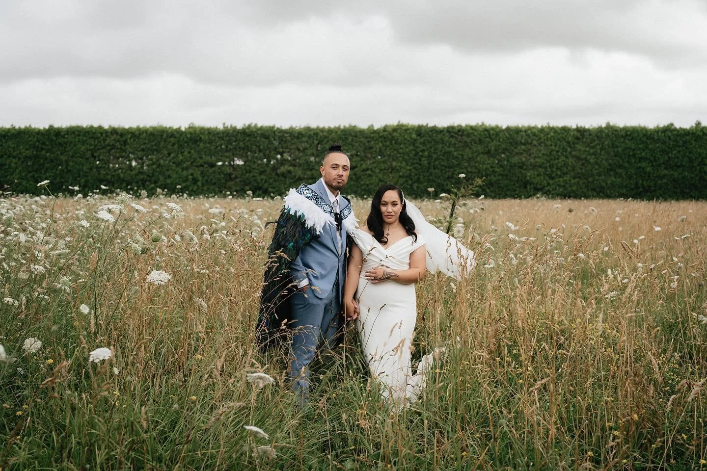A couple dressed in wedding attire standing in a field of tall grass and wildflowers under a cloudy sky, holding hands and facing the camera.