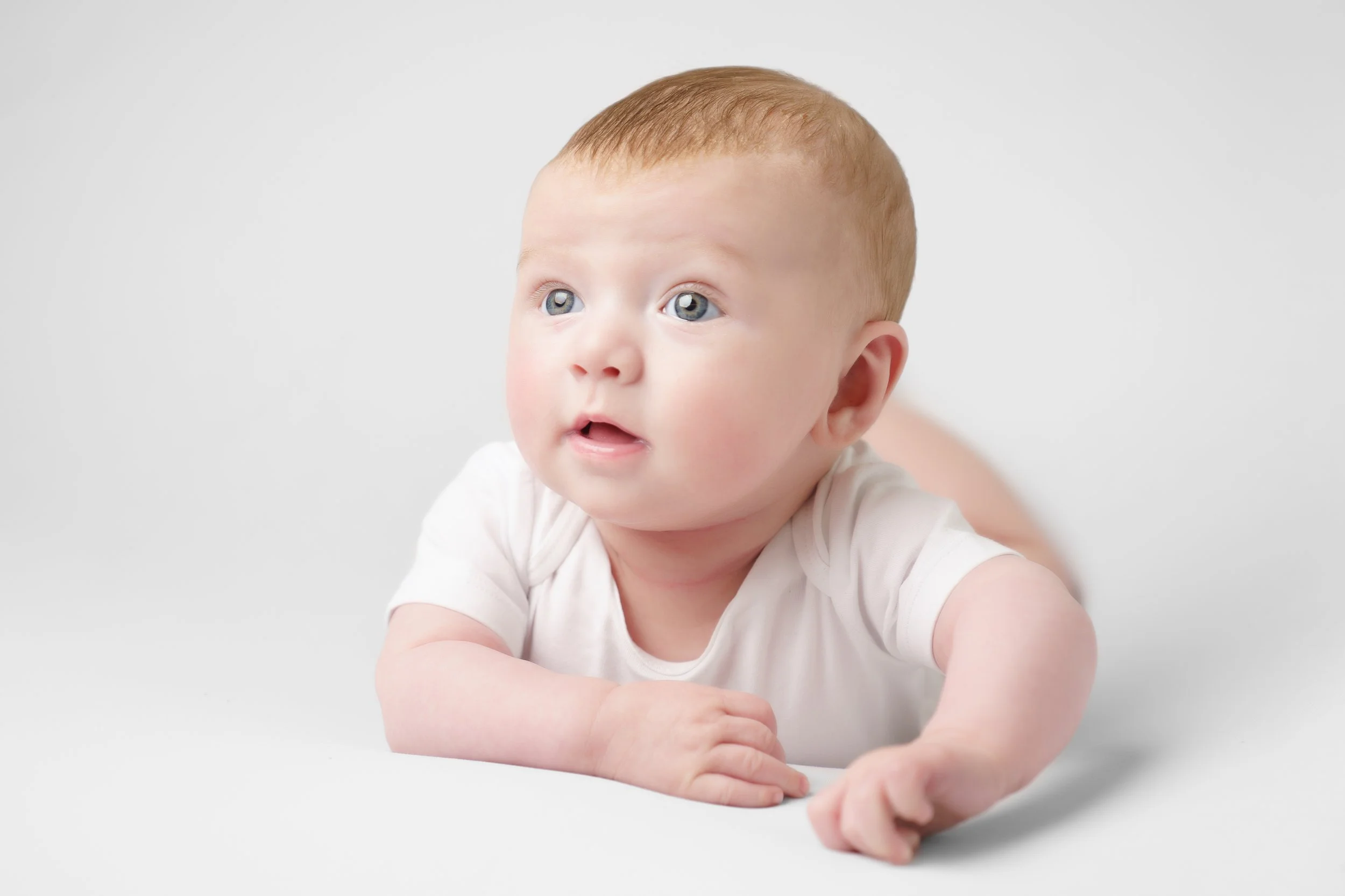 Baby, in witte romper, op de buik, lachend en kijkend in de camera op een witte achtergrond