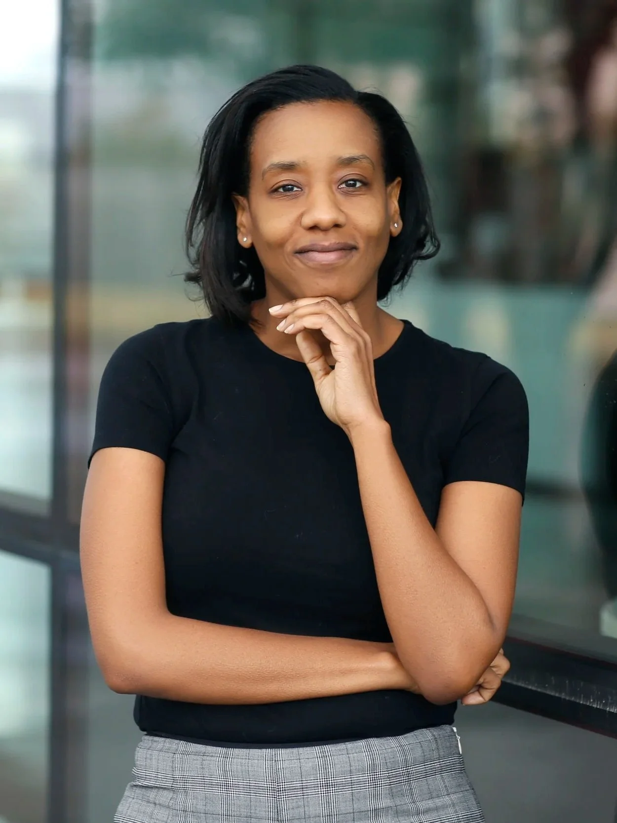 A woman with short black hair wearing a black t-shirt and gray plaid pants, standing with her left arm crossed and her right hand resting on her chin, looking at the camera with a slight smile, in front of a blurred background.