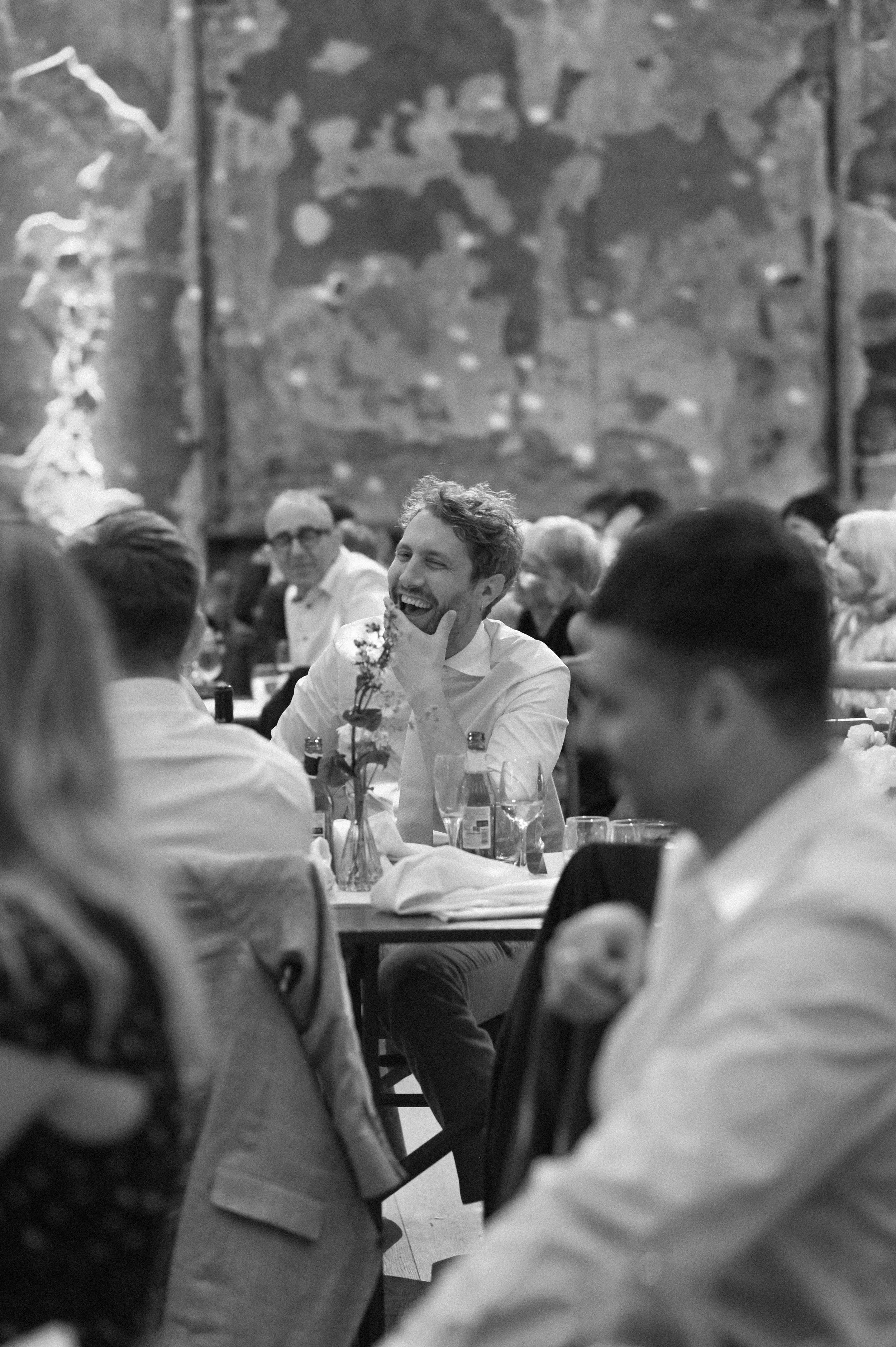 A black and white photograph at Battersea Arts Centre of a white man, a groom on his wedding day, listening to a speech. He is holding his face and laughing.