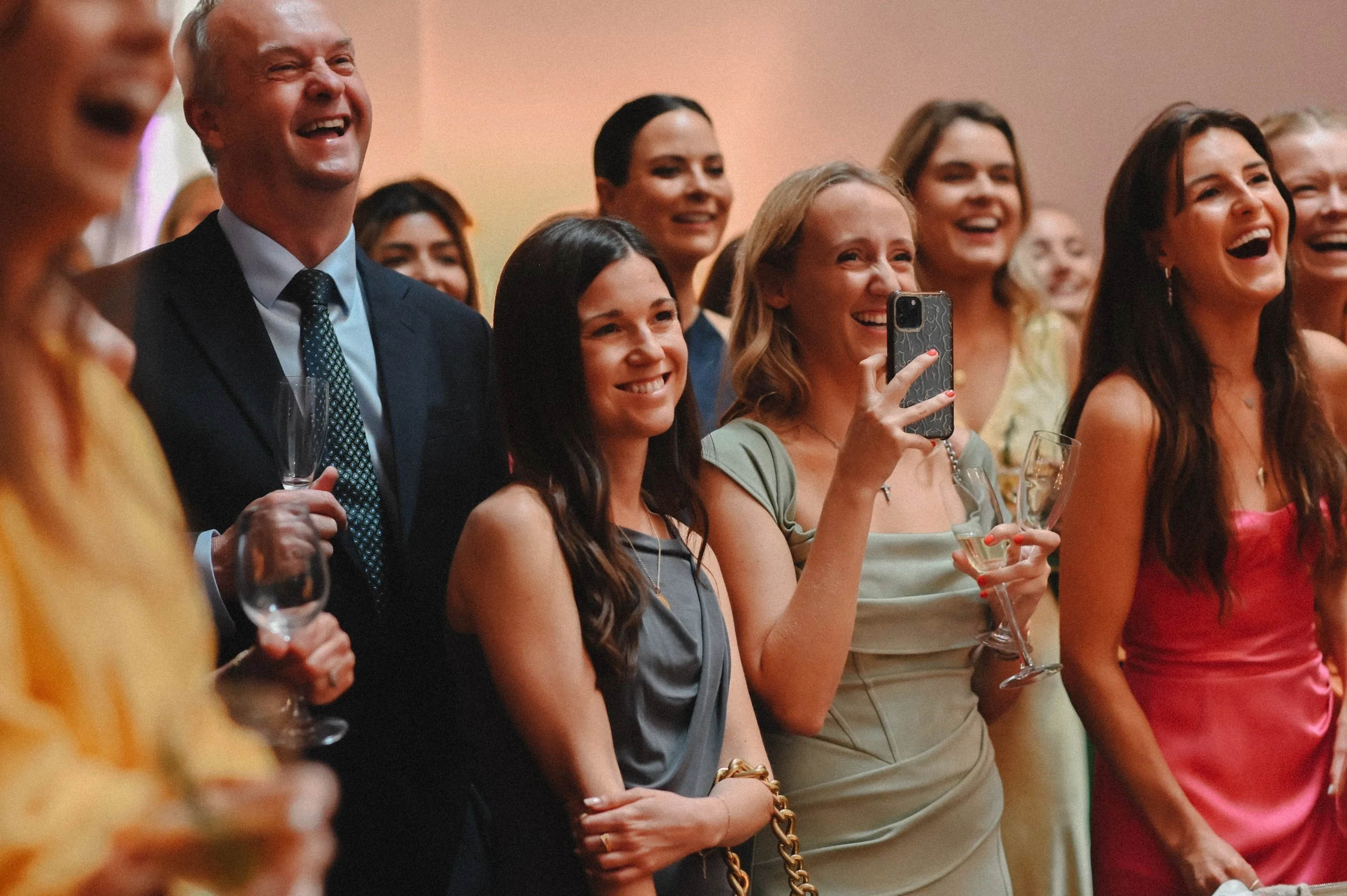 A colour photograph at Oxen Hoath Estate of a group of guests, white men and women, laughing as they watch a speech at a wedding. They are all smiling.