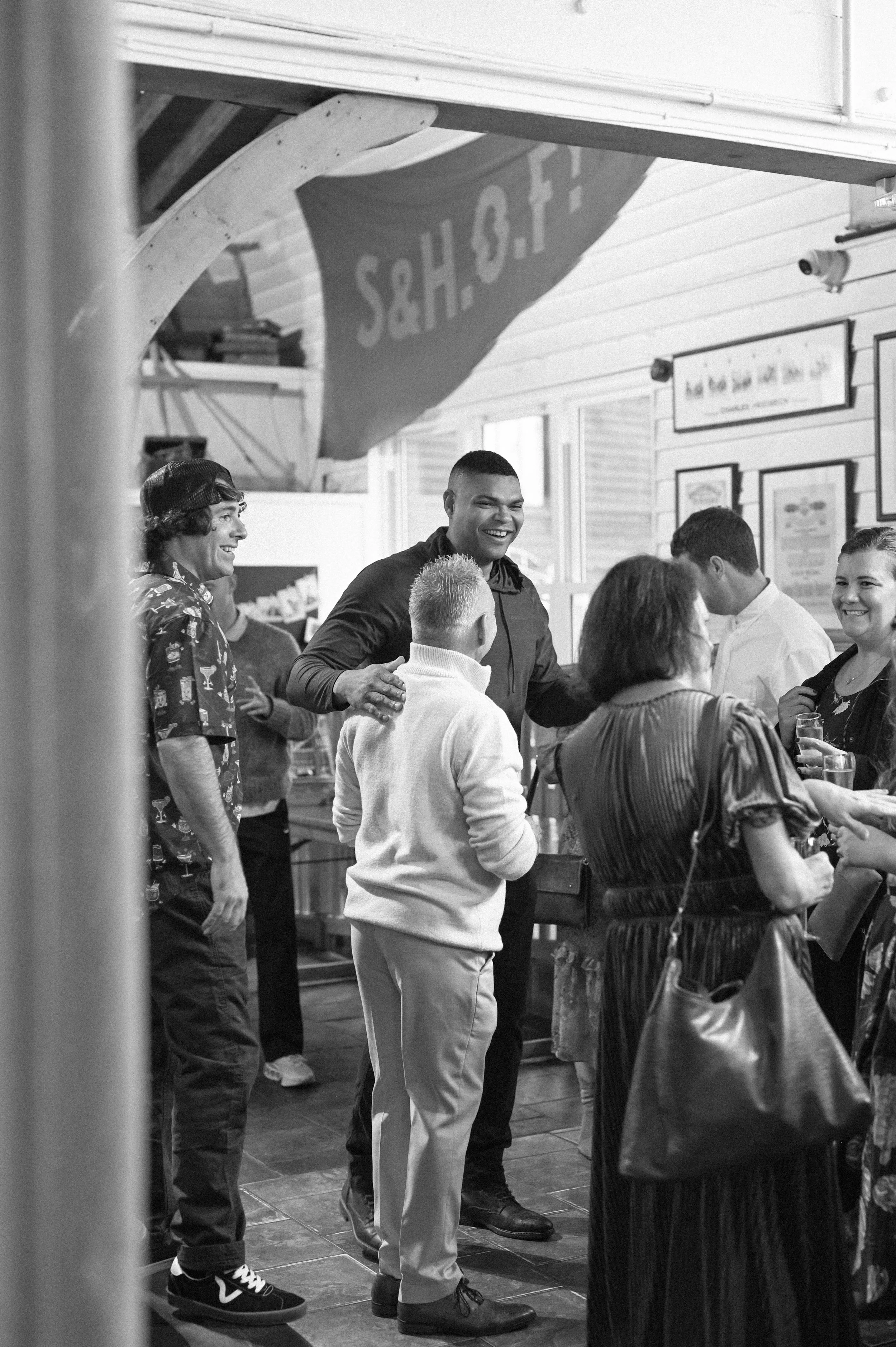 A black and white photograph at East Quay Venue of guests, a Black man and white man, at a wedding embracing and laughing, as the groom, a white man, looks on smiling.