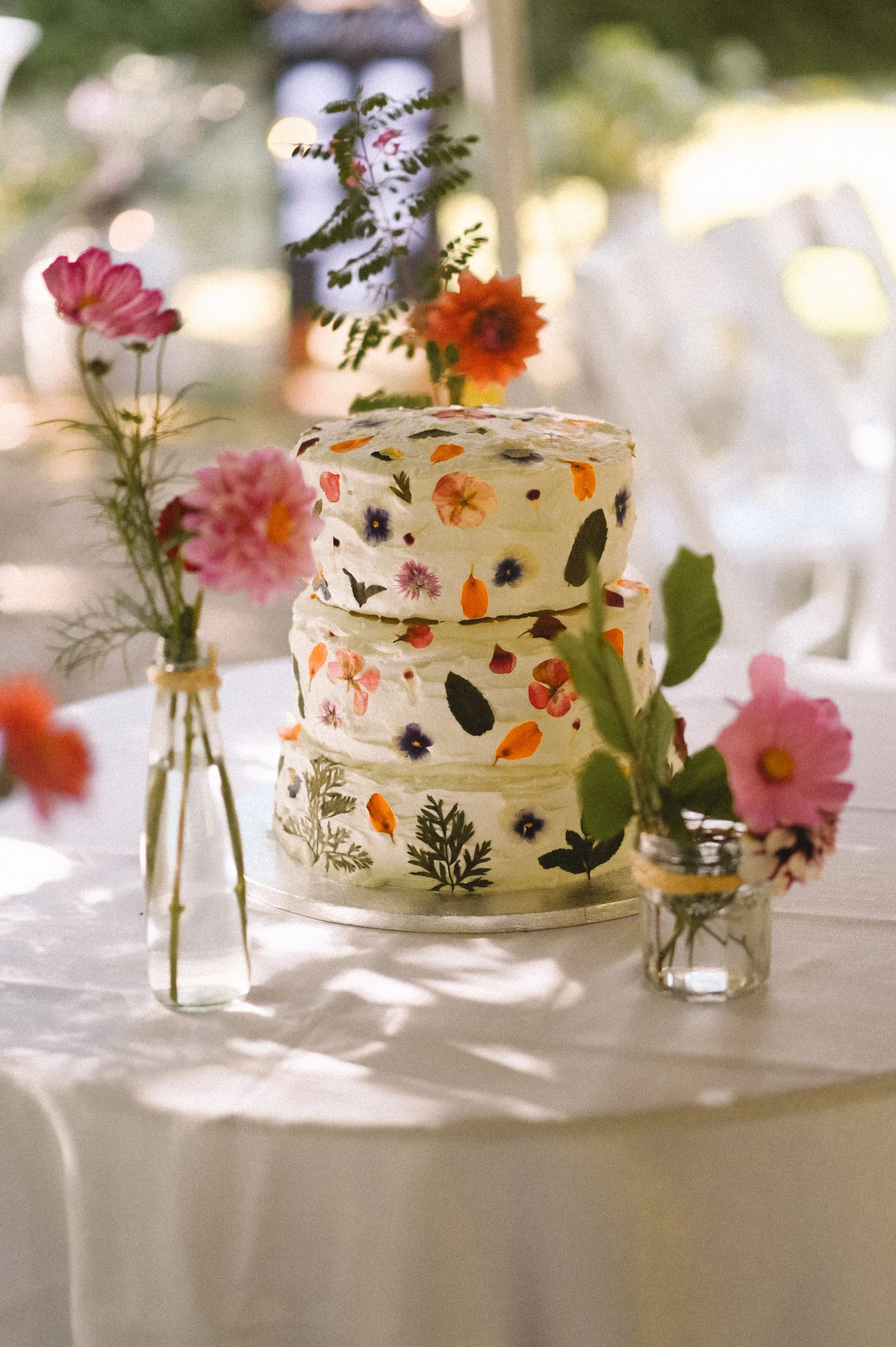 A colour photograph at Sausmarez Manor of a three tiered cake, covered in different real flowers. There is dappled light across the table.