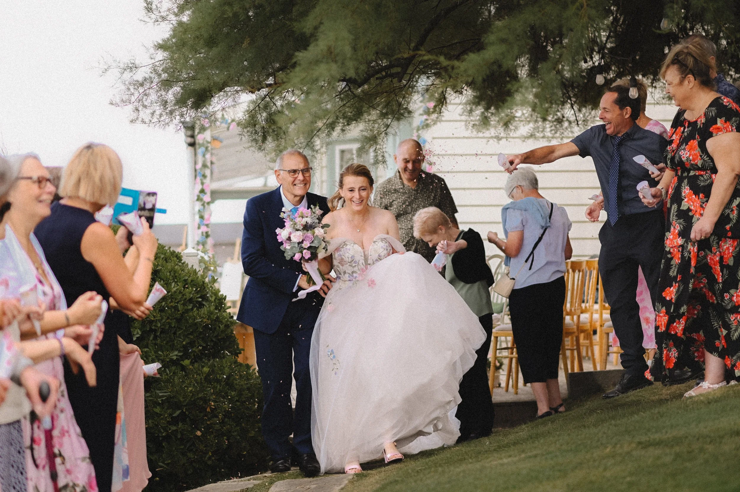 A colour photograph of a white man and woman, a bride and groom at Beacon House on their wedding day, beginning their confetti walk. The bride is wearing a pink dress embroidered with flowers and the groom is in a blue suit. They are both laughing.