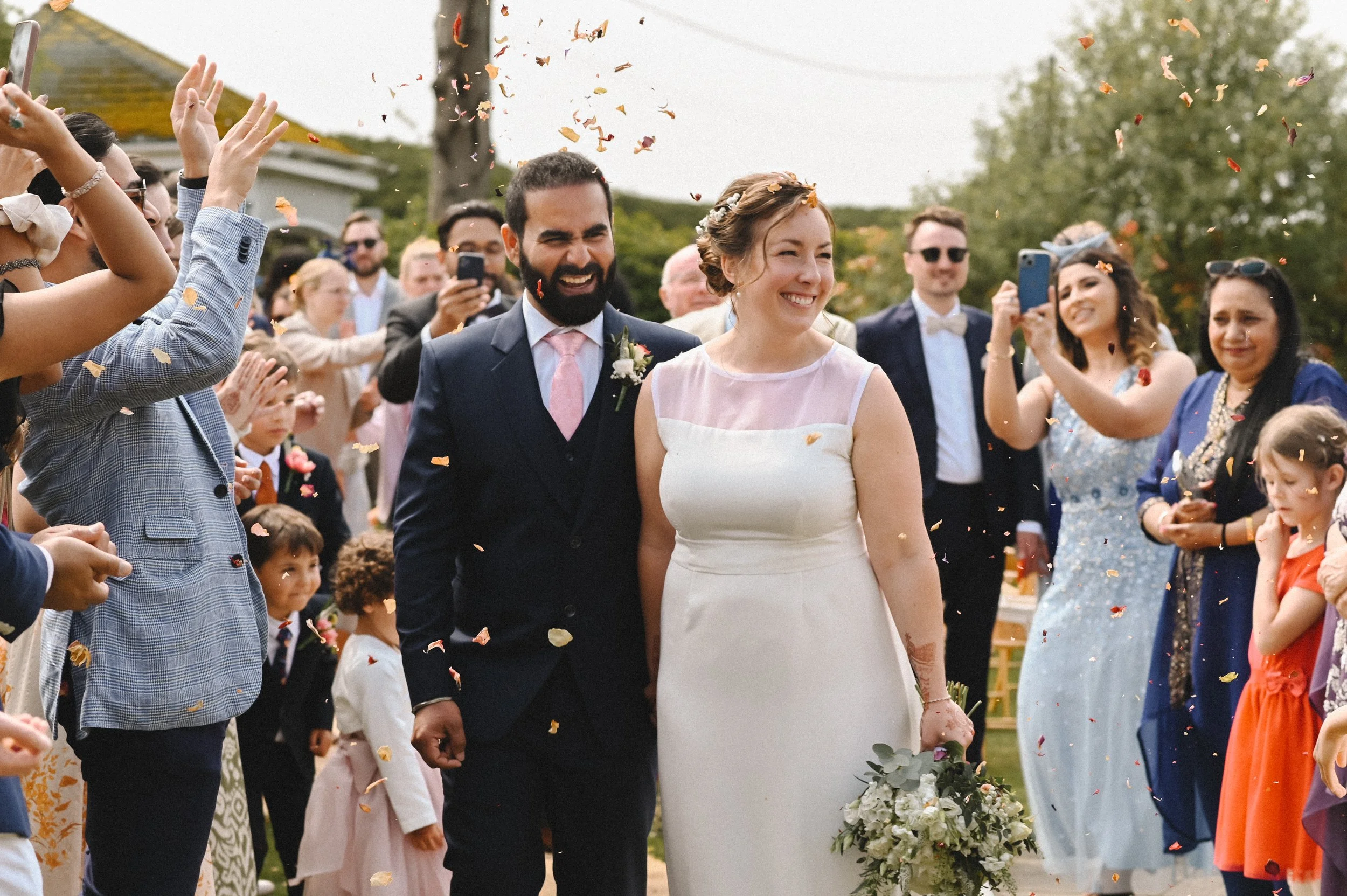 A colour photograph at the Crescent Turner Hotel of a couple, a Brown man and white woman, on their wedding day, smiling from ear to ear as confetti is thrown around them. The photograph feels joyful.