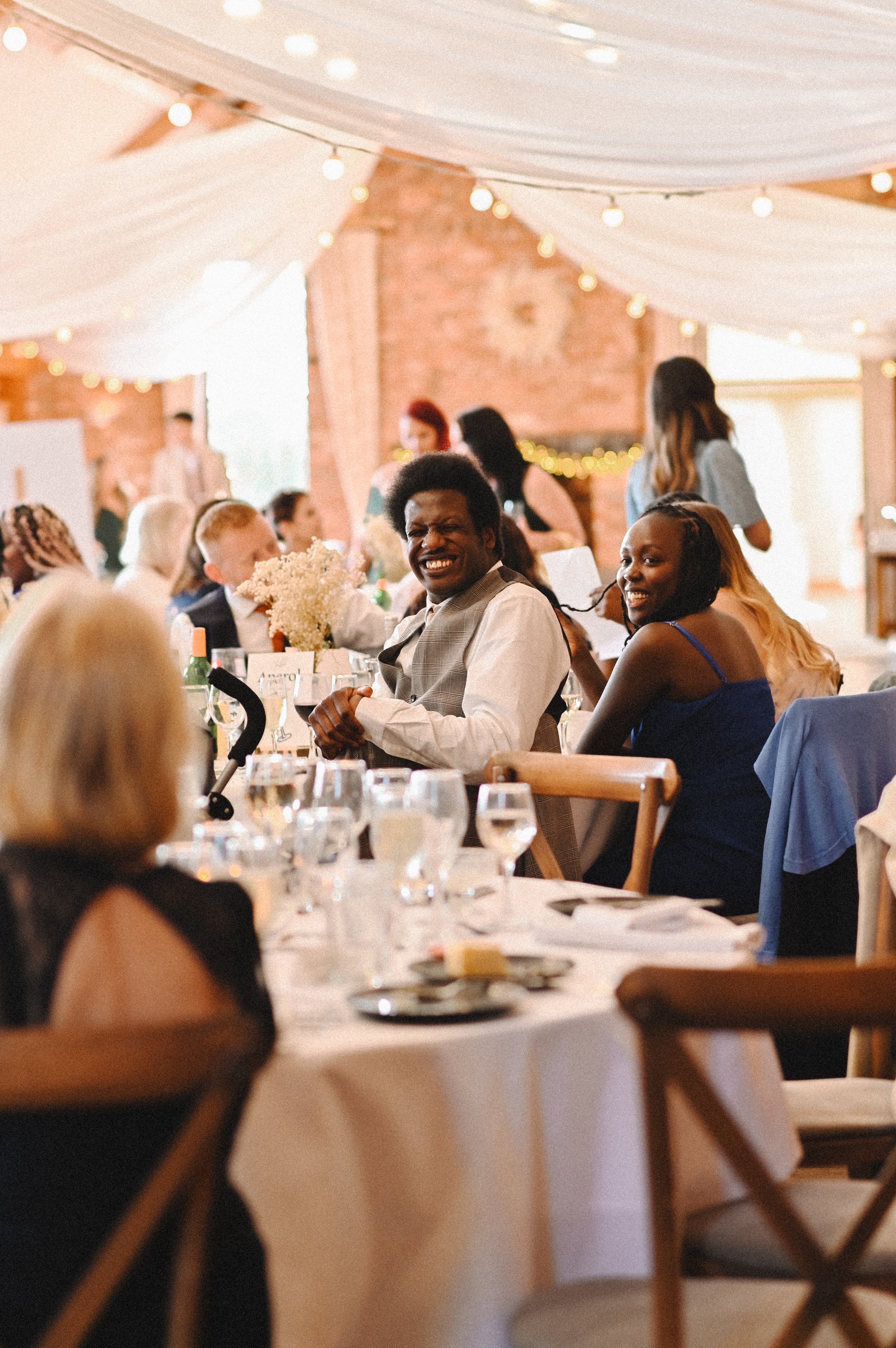 A colour photograph of a Black man and woman, guests at a wedding at Westerham Golf Club. They are smiling and laughing as they speak to another guest in the foreground.
