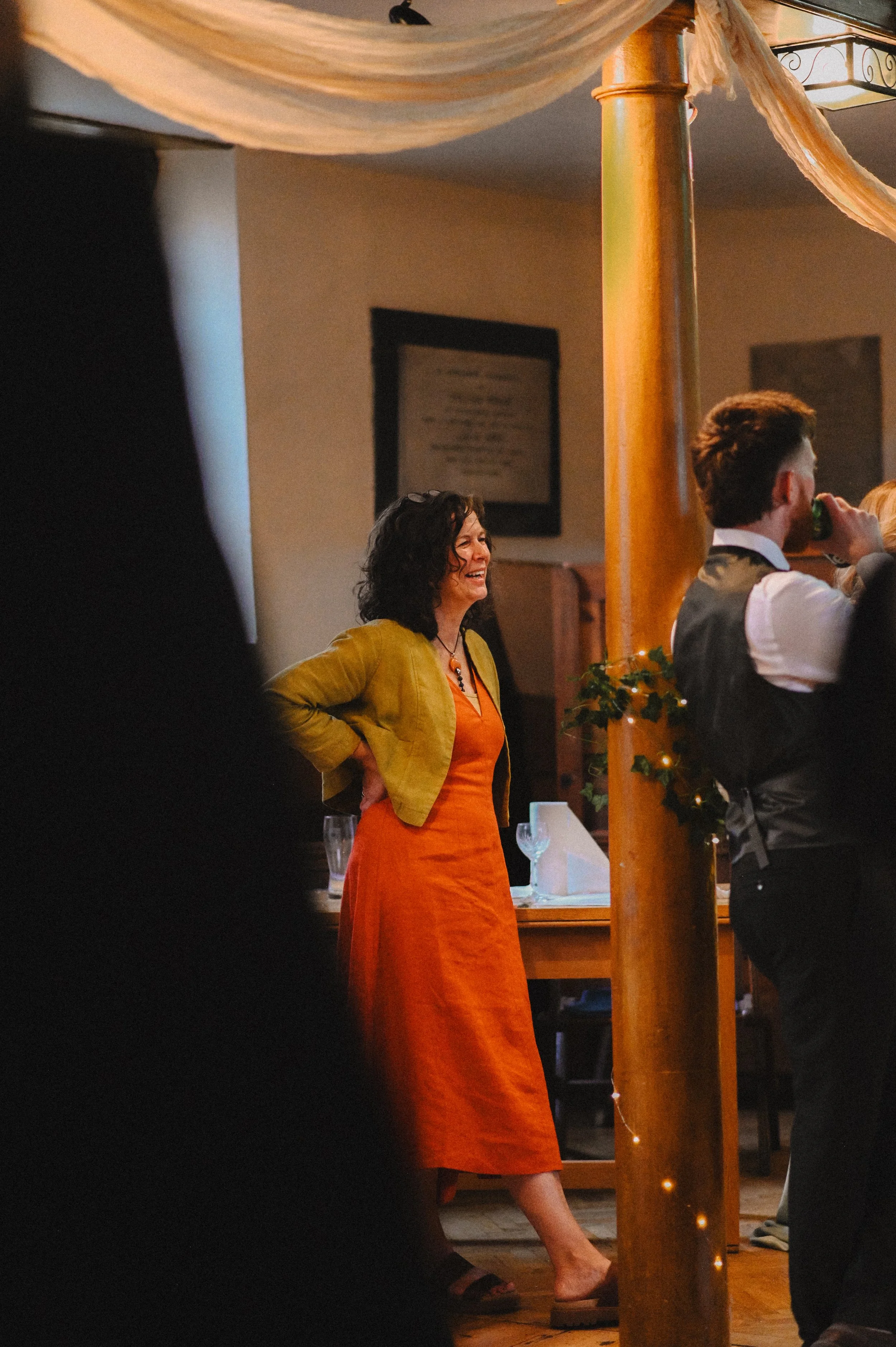 A colour photograph of a white woman, the mother of a groom watching as speeches are made at his wedding. She is laughing.