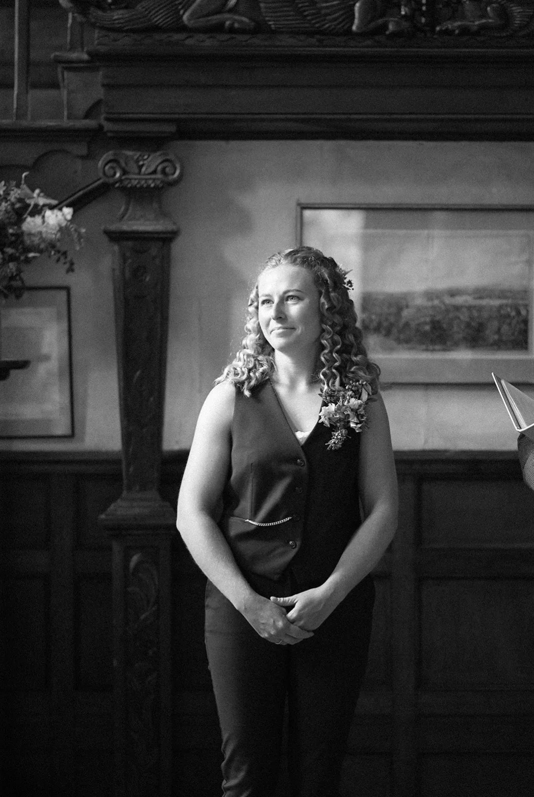 A black and white photograph at Sausmarez Manor of a white woman, a bride seeing her bride come down the stairs into their wedding ceremony. She is wearing a waistcoat suit and smiling with adoration.