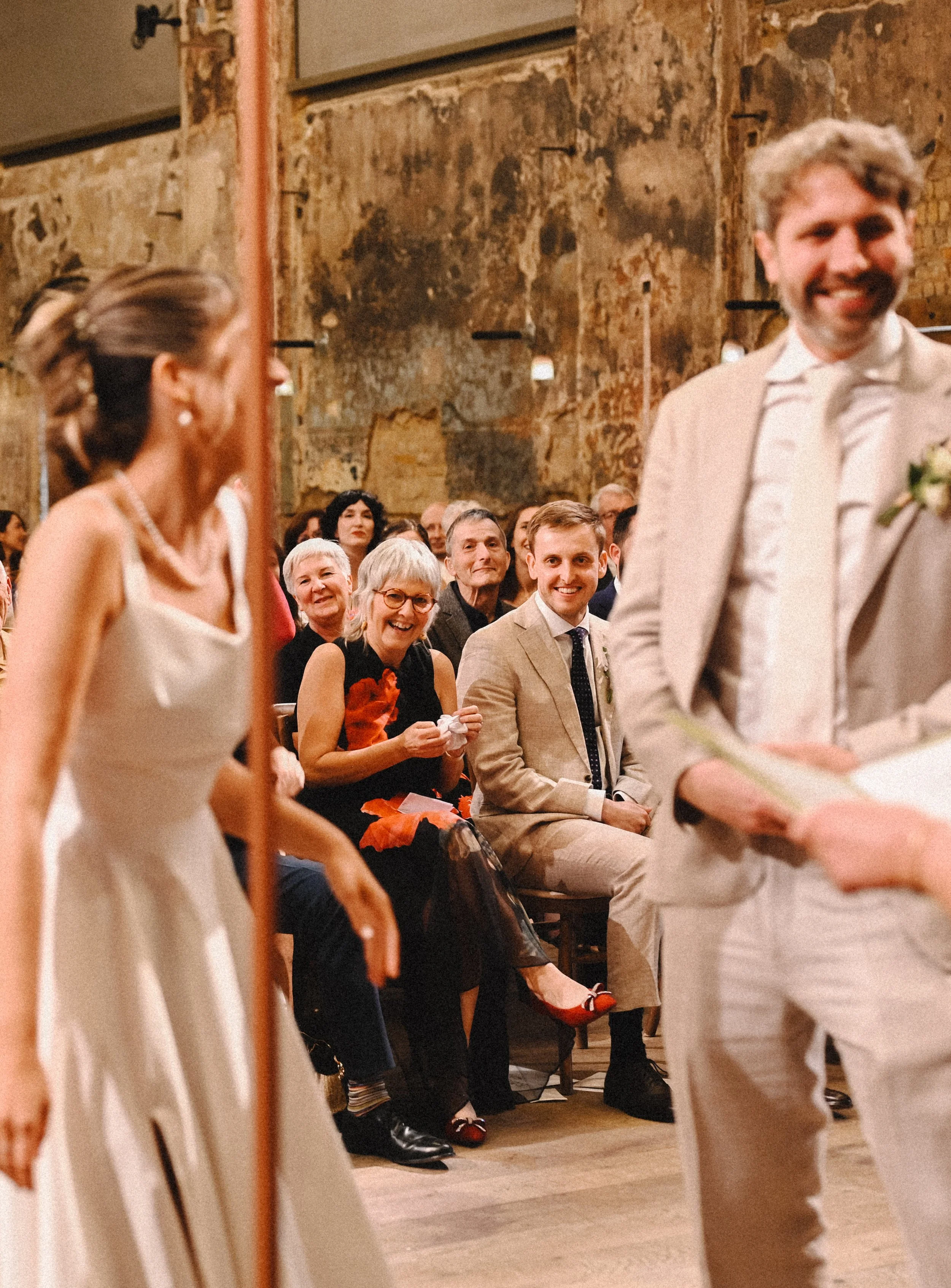 A colour photograph at Battersea Arts Centre of a bride's mother and brother, a white man and woman, laughing as they watch their daughter and sister, and her groom giggling during their ceremony.