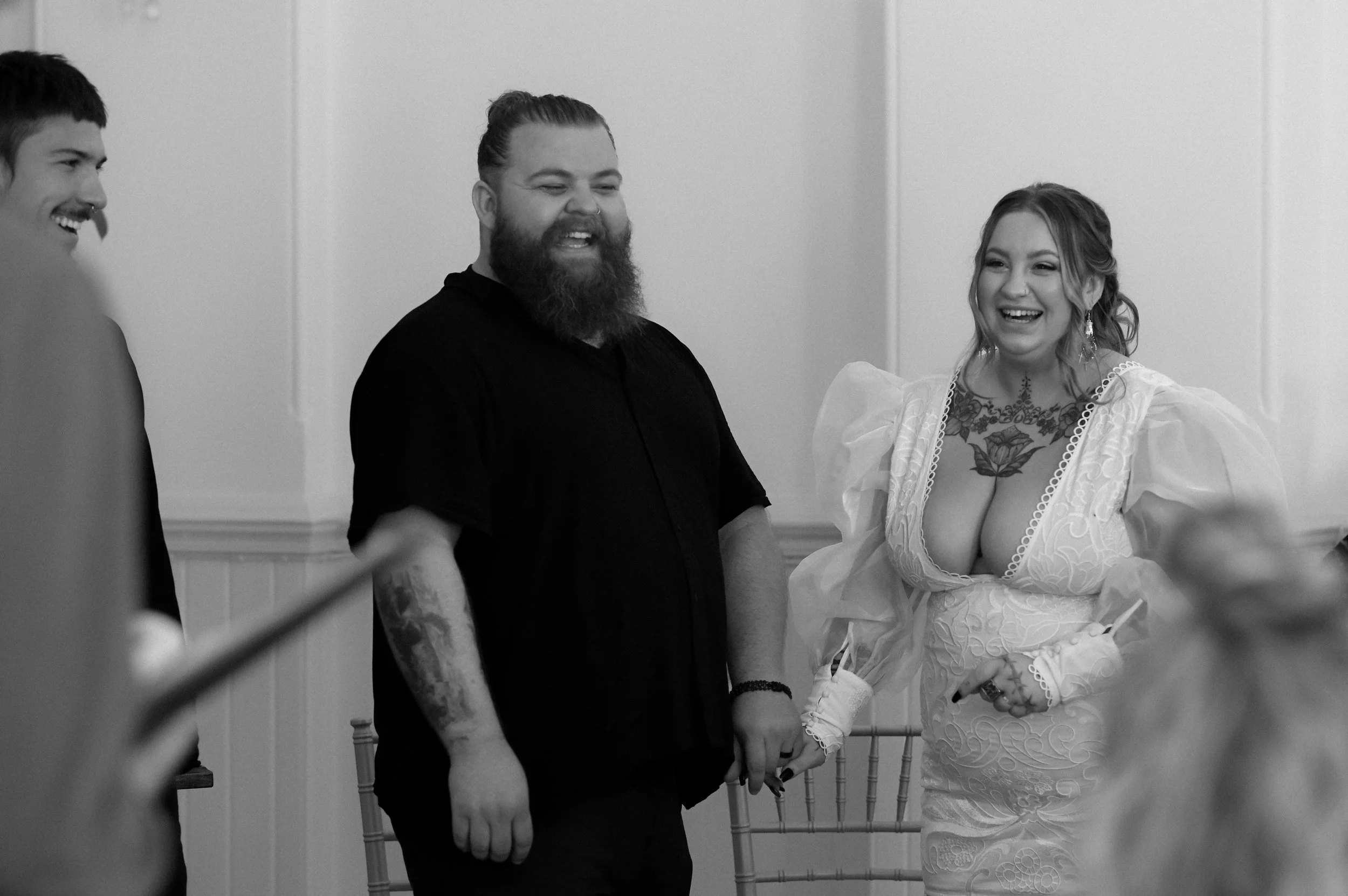 A black and white photograph at Brighton Town Hall of a bride and groom, a white man and woman, during their wedding ceremony. They are holding hands and laughing. They both have tattoos.