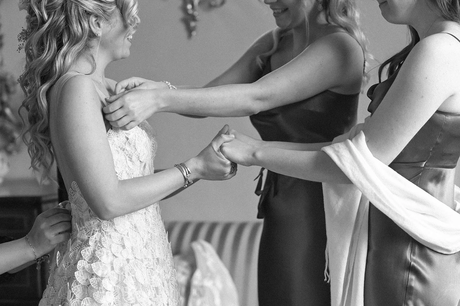 A black and white photograph at Chenies Manor of a white woman, a bride getting ready for her wedding. There are pairs of hands doing up her dress, adjusting her collar and holding her hands.