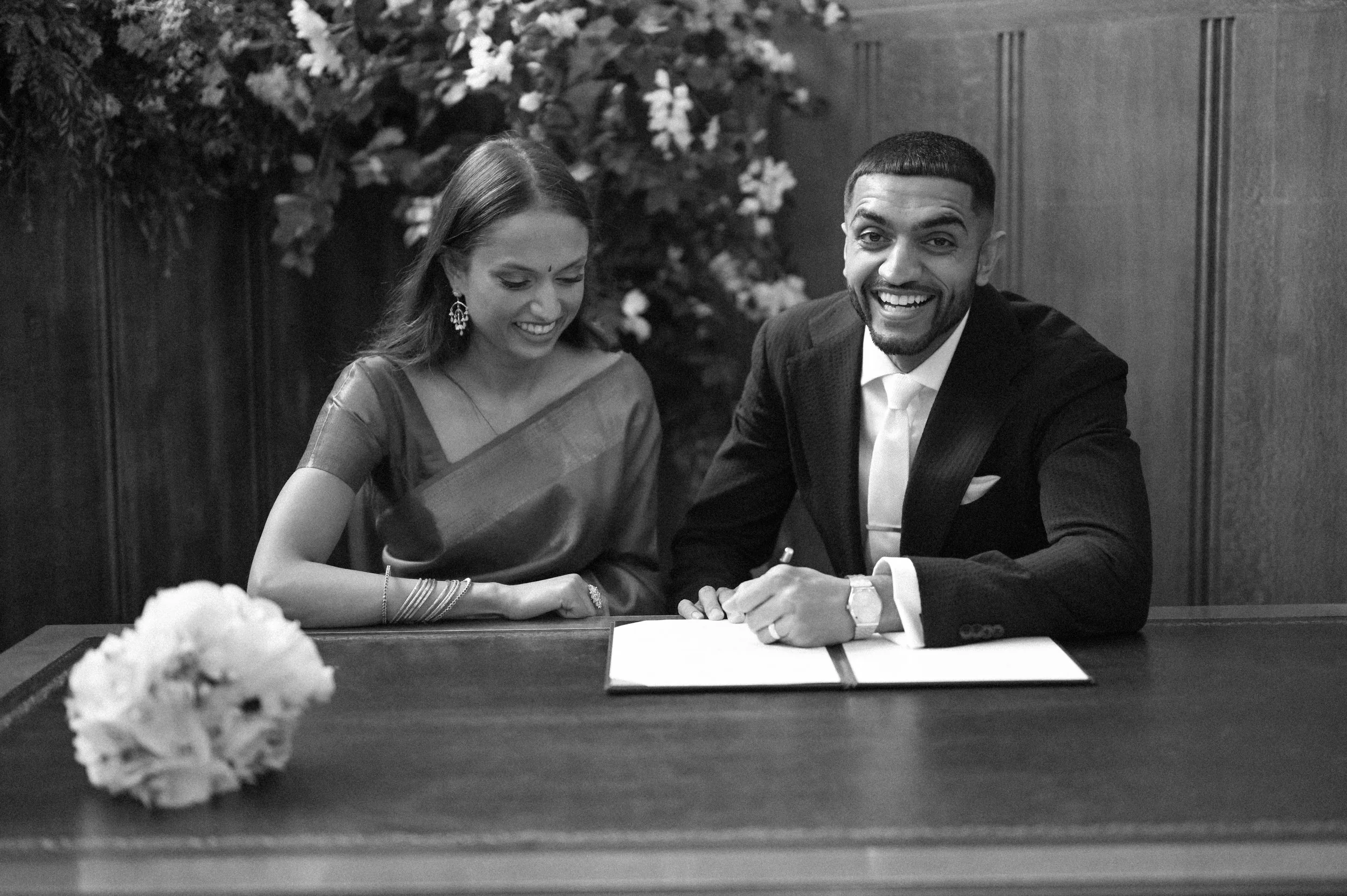 A black and white photograph at Kingston Town Hall of a Brown man and woman, a bride and groom signing their marriage certificate. The groom is smiling at the camera, and the bride is wearing a bridal sari and grinning and looking down.