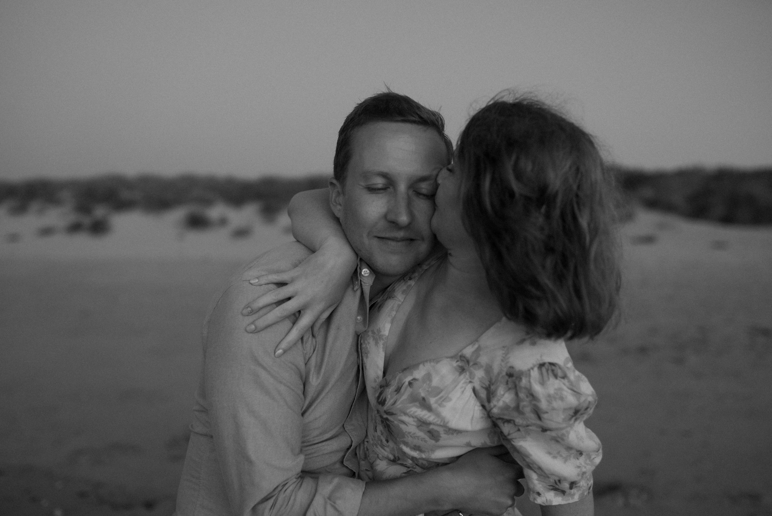 A black and white photograph at Port Soif beach, of a white man and woman, a couple embracing. The woman is kissing her husband's face and he smiles as his eyes are closed.