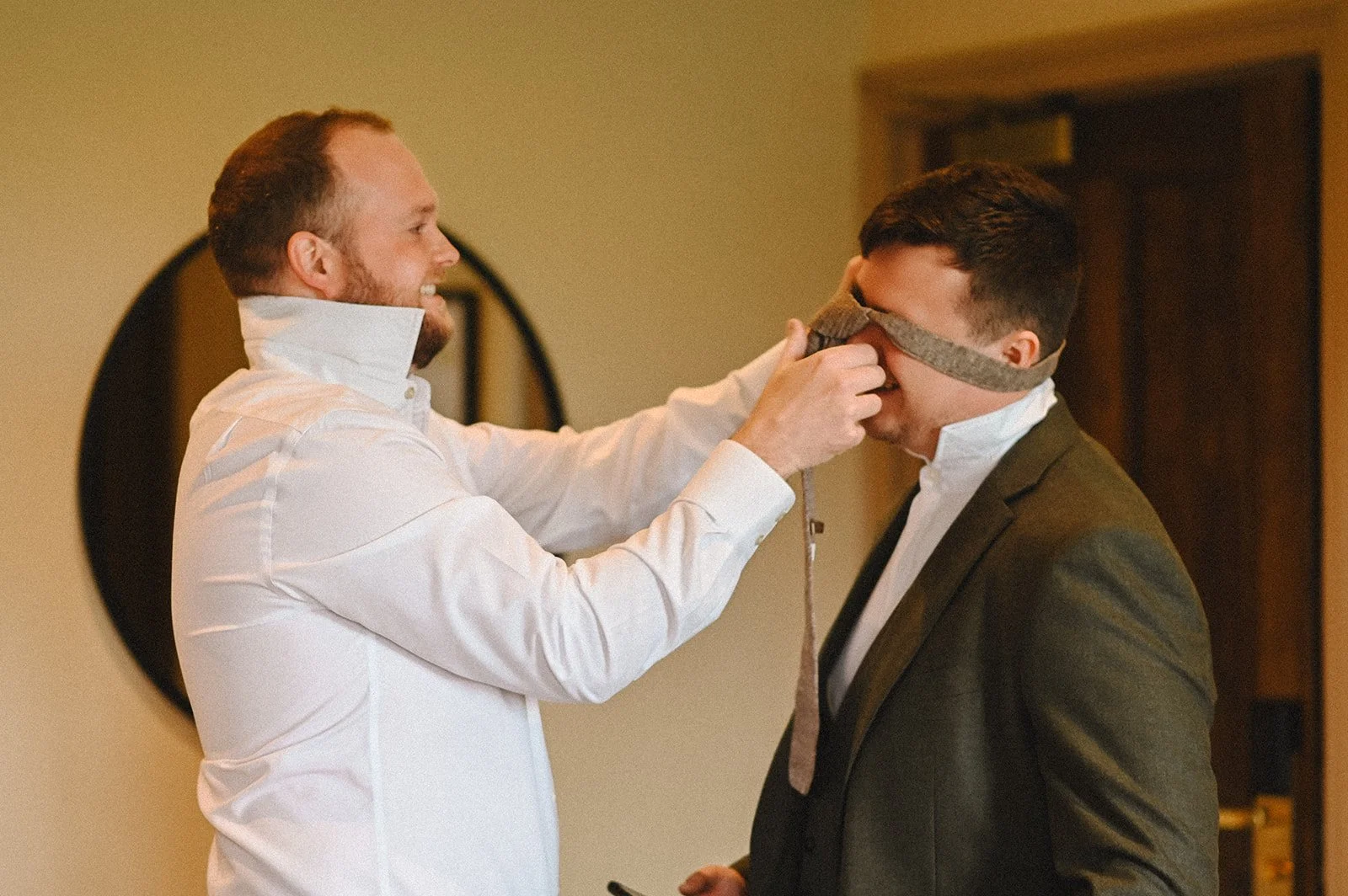 A colour photograph at Chenies Manor of two white men, a groom having his tie put on by his best man. It is stuck on his head, and they are both laughing.