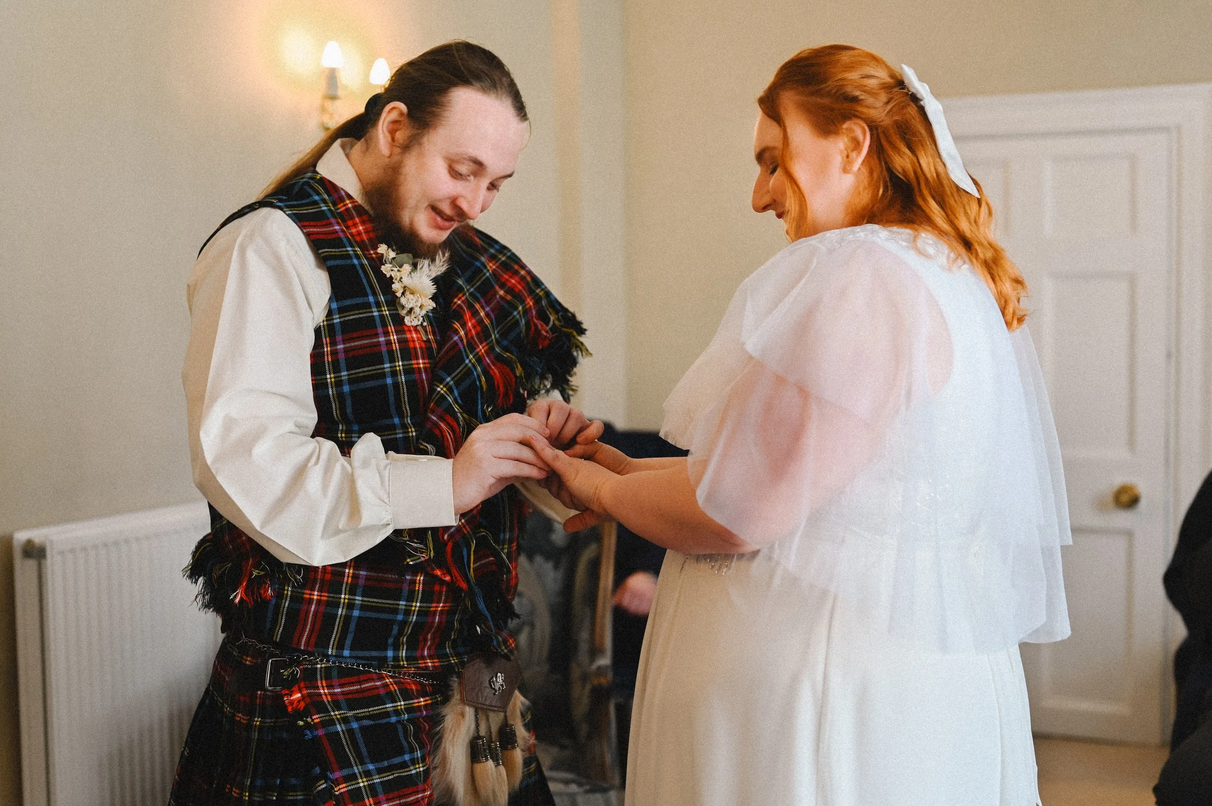A colour photograph at Canterbury Town Hall of a bride and groom, a white man and woman, exchanging rings at their wedding. They are both smiling from ear to ear. The groom is wearing traditional Scottish dress.