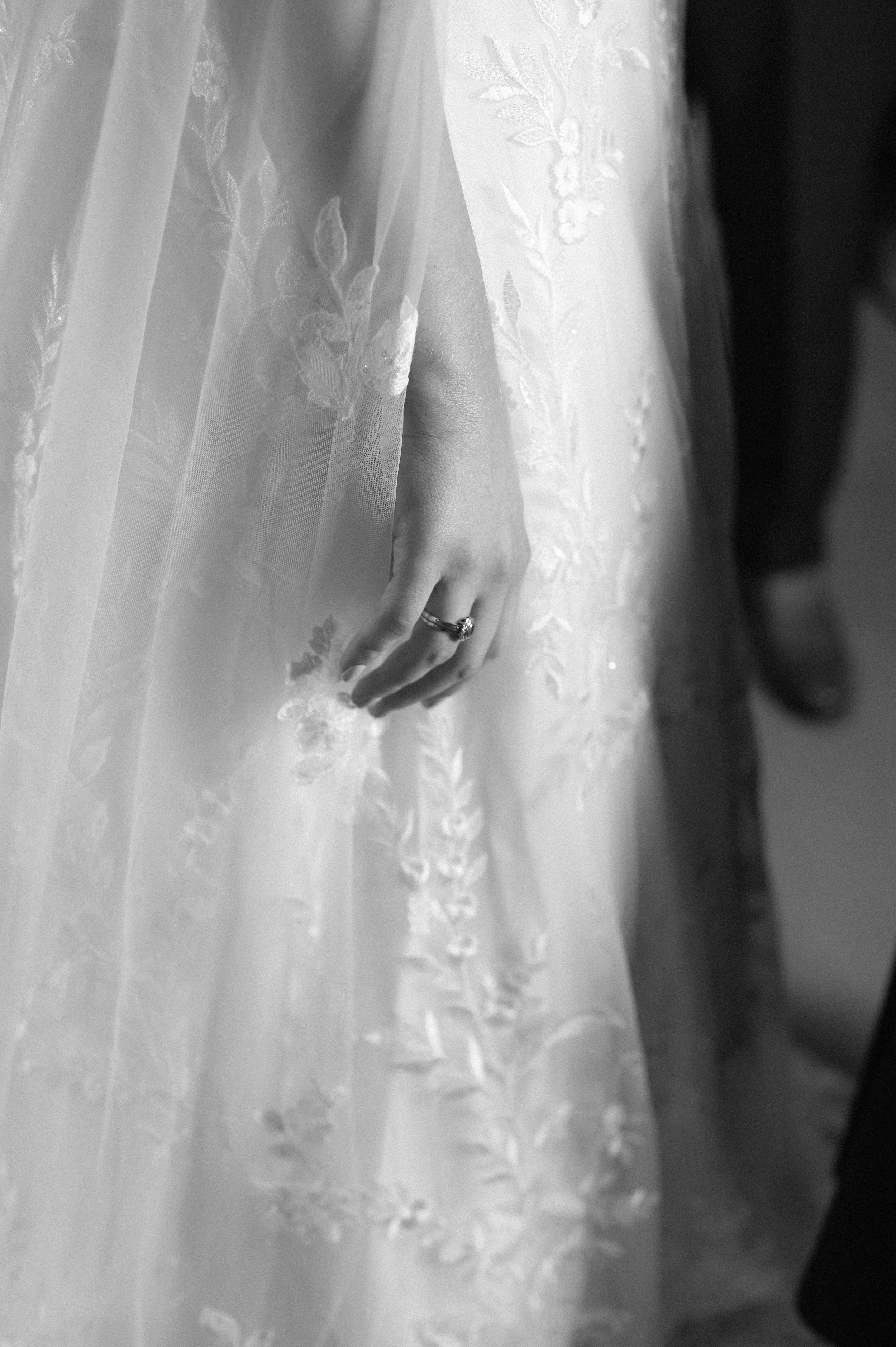 A black and white photograph at The George and Dragon of a white woman, a bride's hand wrapped in her veil as she gets ready to leave for her wedding. The details in her dress and her engagement ring can be seen.
