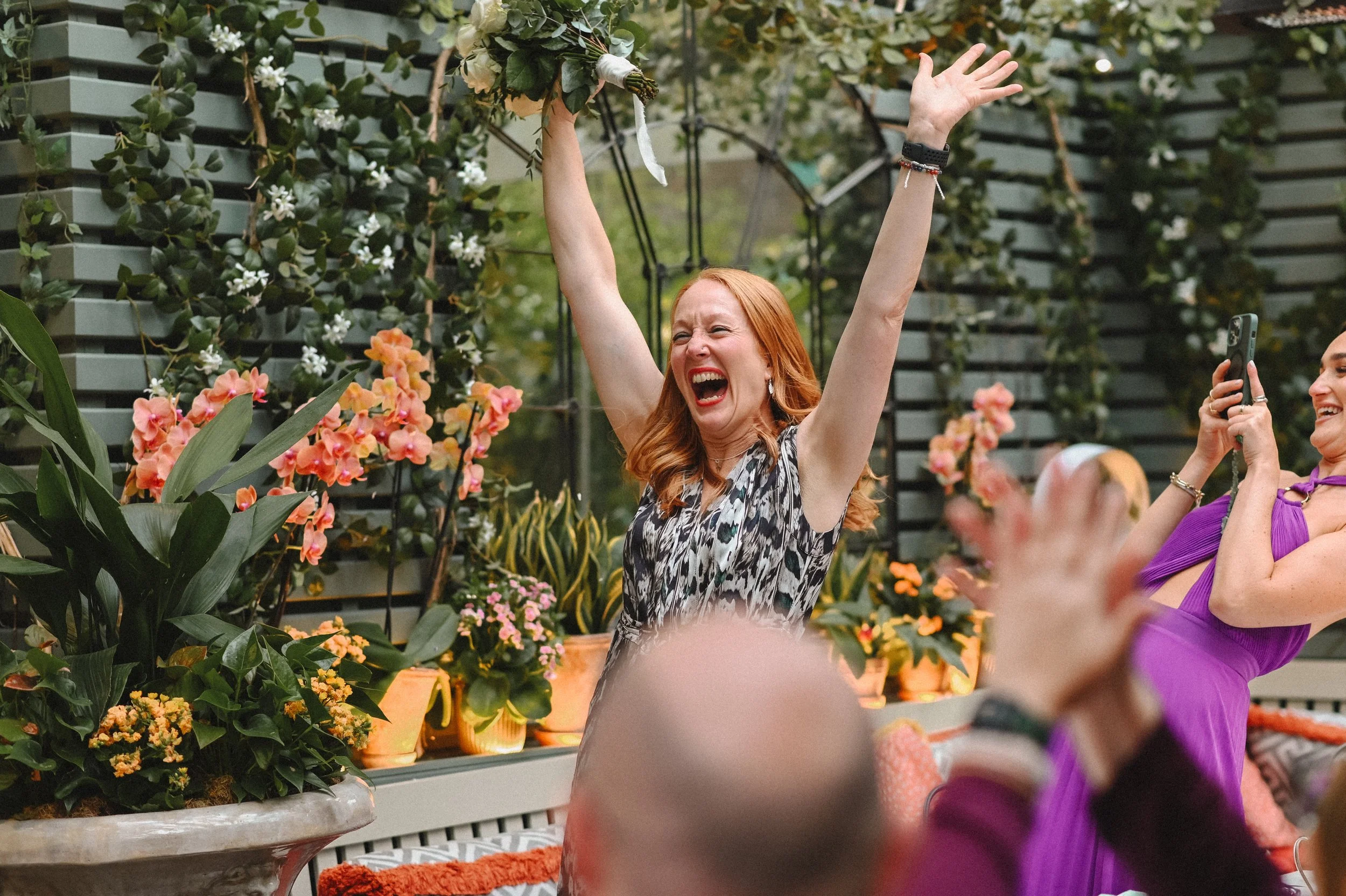 A colour photograph at The Ivy Chelsea of a white woman, a guest at a wedding. She has just caught the bride's bouquet and is holding it above her head and celebrating.