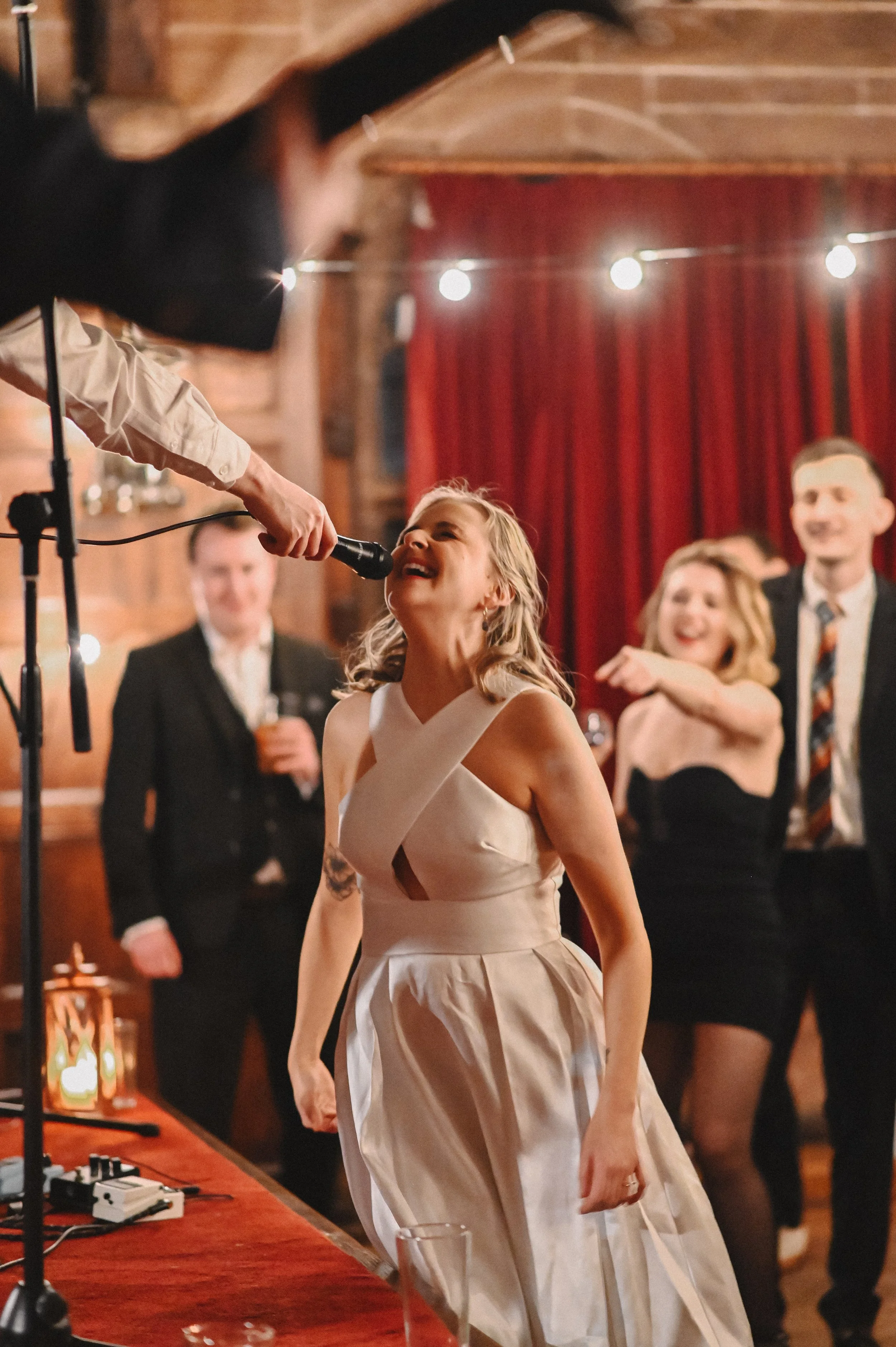 A colour photograph of white woman, a bride at her wedding reception singing into a microphone being held out by her new husband from stage. She is being cheered on in the background by guests.