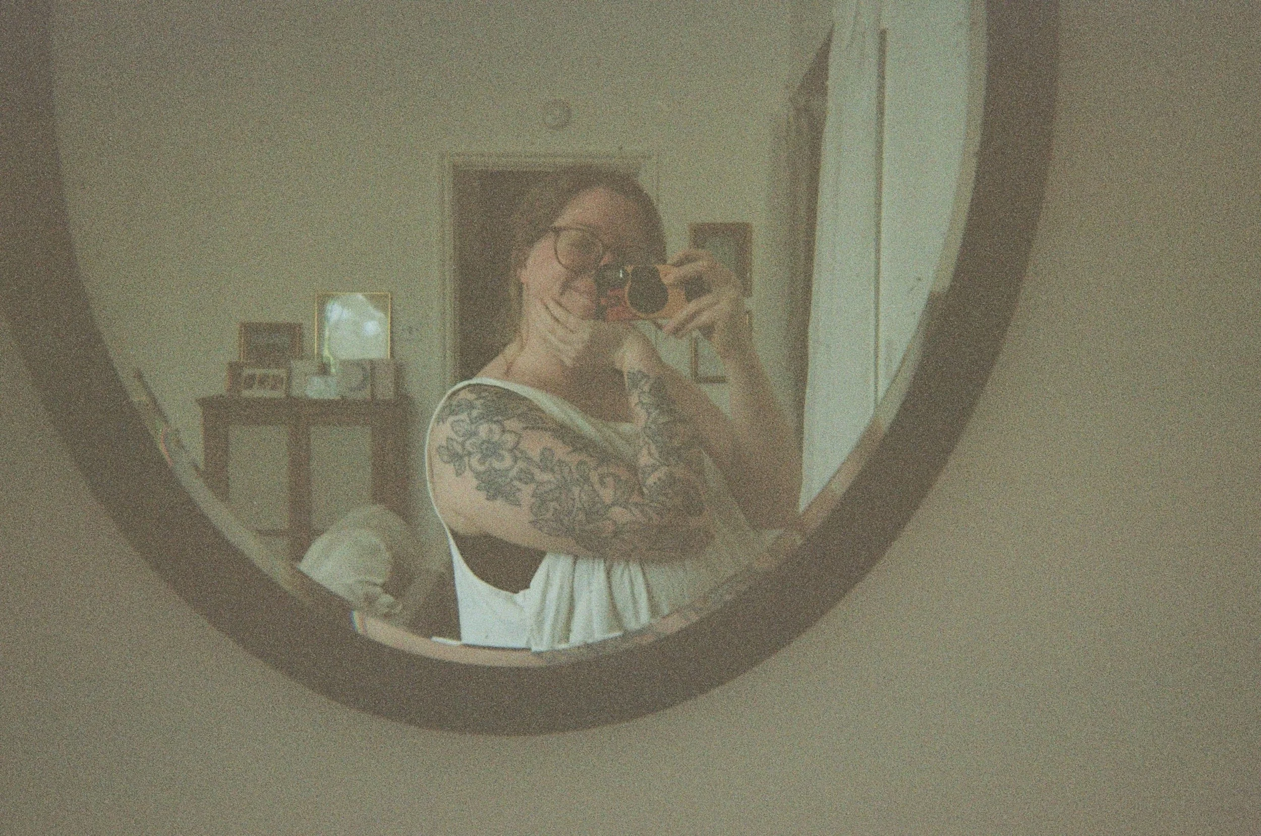 A scanned colour photograph of a white woman taking the picture with a film camera while she smiles in a mirror with her eyes closed. She has glasses and a full arm of floral tattoos.