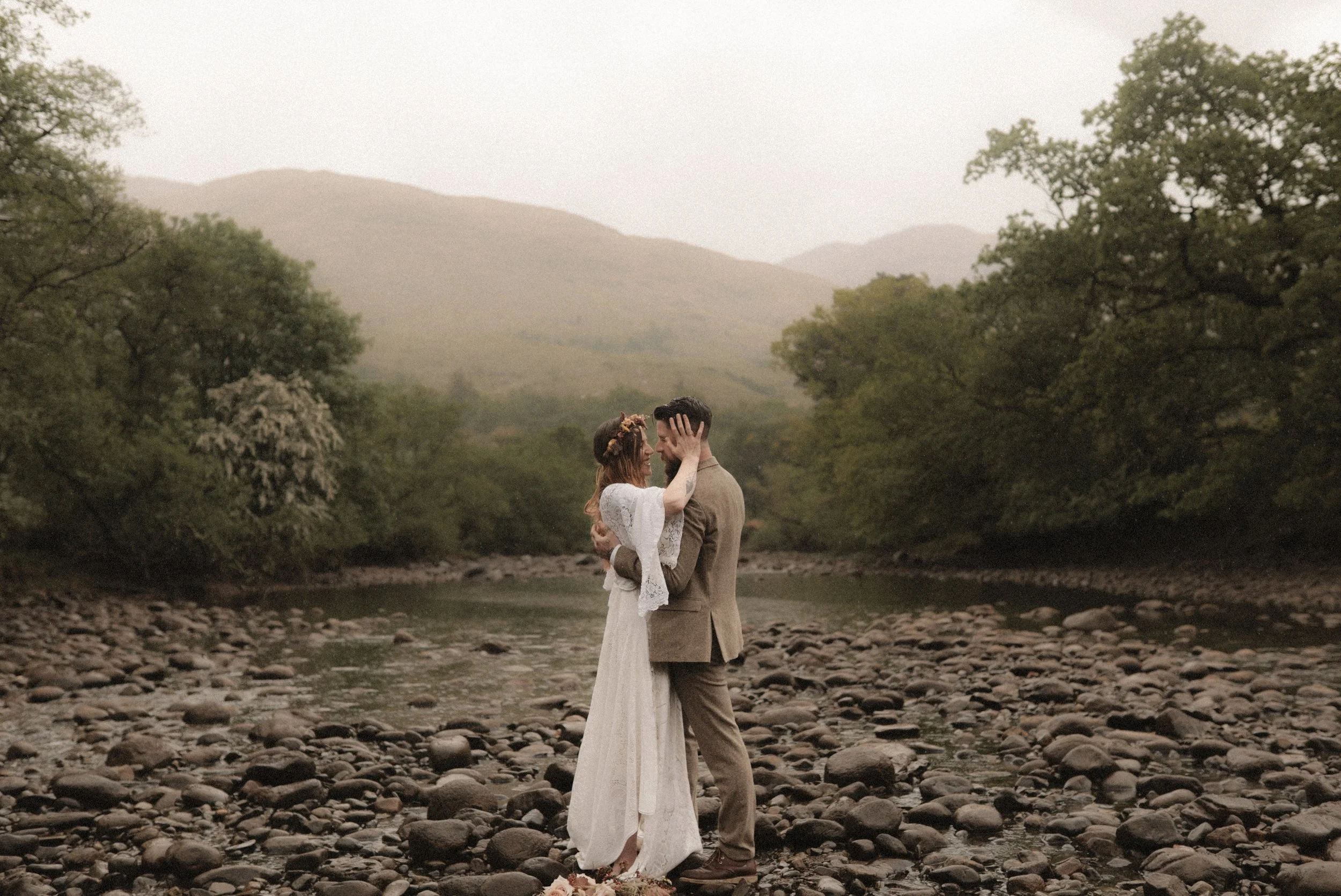 A colour photograph in the Scottish Highlands of a white man and woman, a bride and groom during their elopement, embracing in the middle of a river. The bride is holding her groom's face as they both smile.