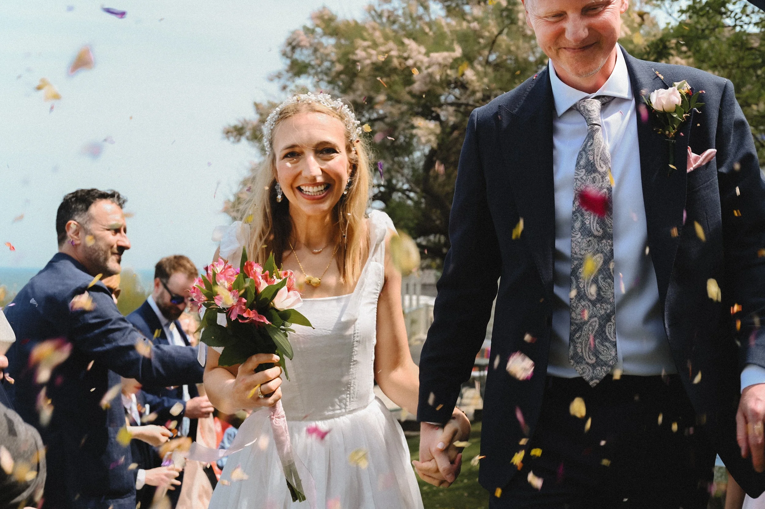 A colour photograph at The Beacon House of a white couple walking through confetti petals. They are holding hands and smiling from ear to ear. The groom is wearing a blue suit and patterned tie, and the bride is wearing a white corset dress.