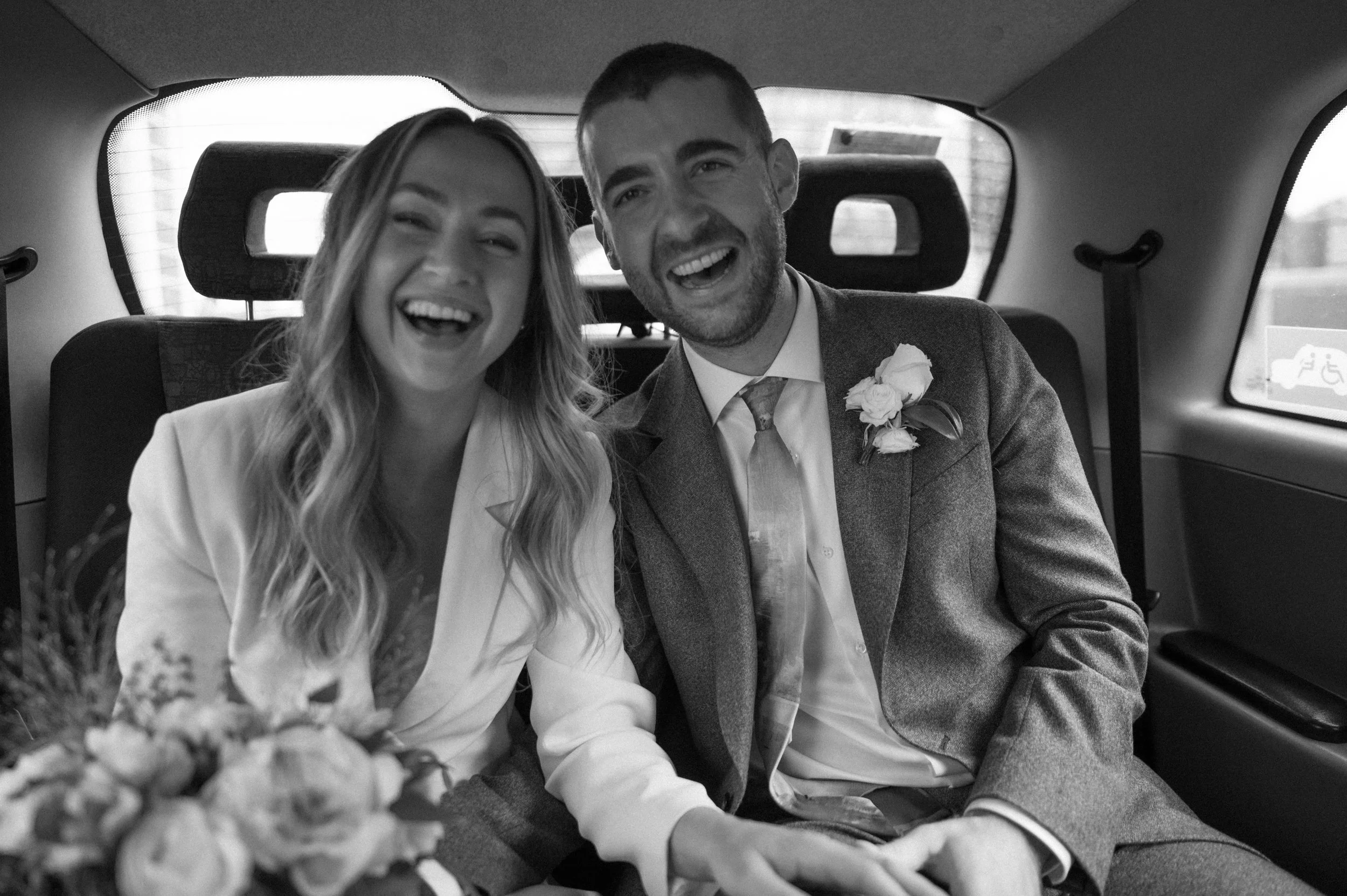 A black and white photograph in London of a white man and woman, a couple in the back of a cab on the way to their wedding reception. They are holding hands and laughing at the camera. They are both wearing suits.
