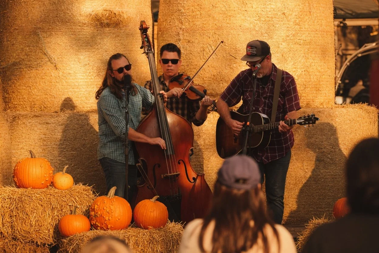 Three musicians playing instruments on stage with autumn pumpkins and hay bales in the background, at an outdoor fall event.