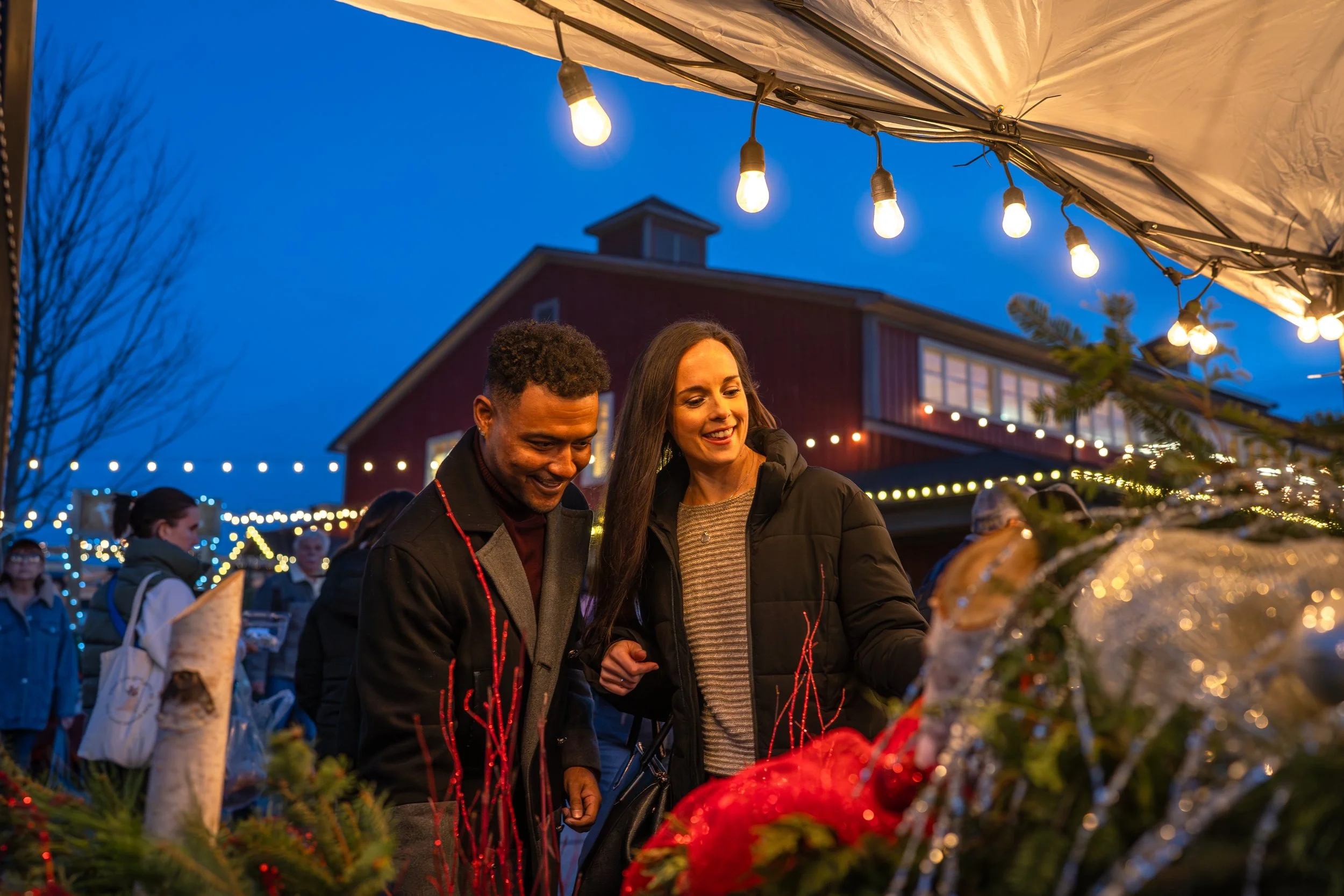 People shopping outside at St. Jacobs Farmers Market during the winter holiday season, decorated with Christmas trees and string lights.