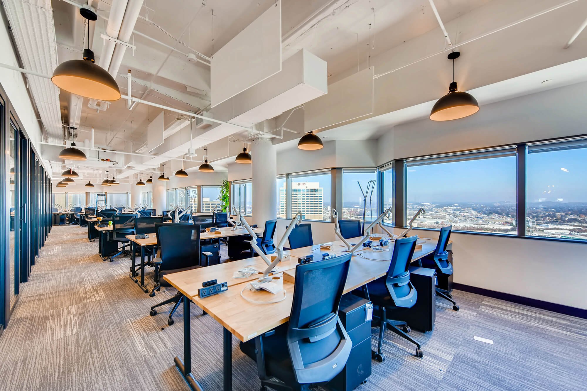  Modern office interior featuring long rows of desks with blue office chairs, hanging pendant lights, large windows, and an urban cityscape view. 