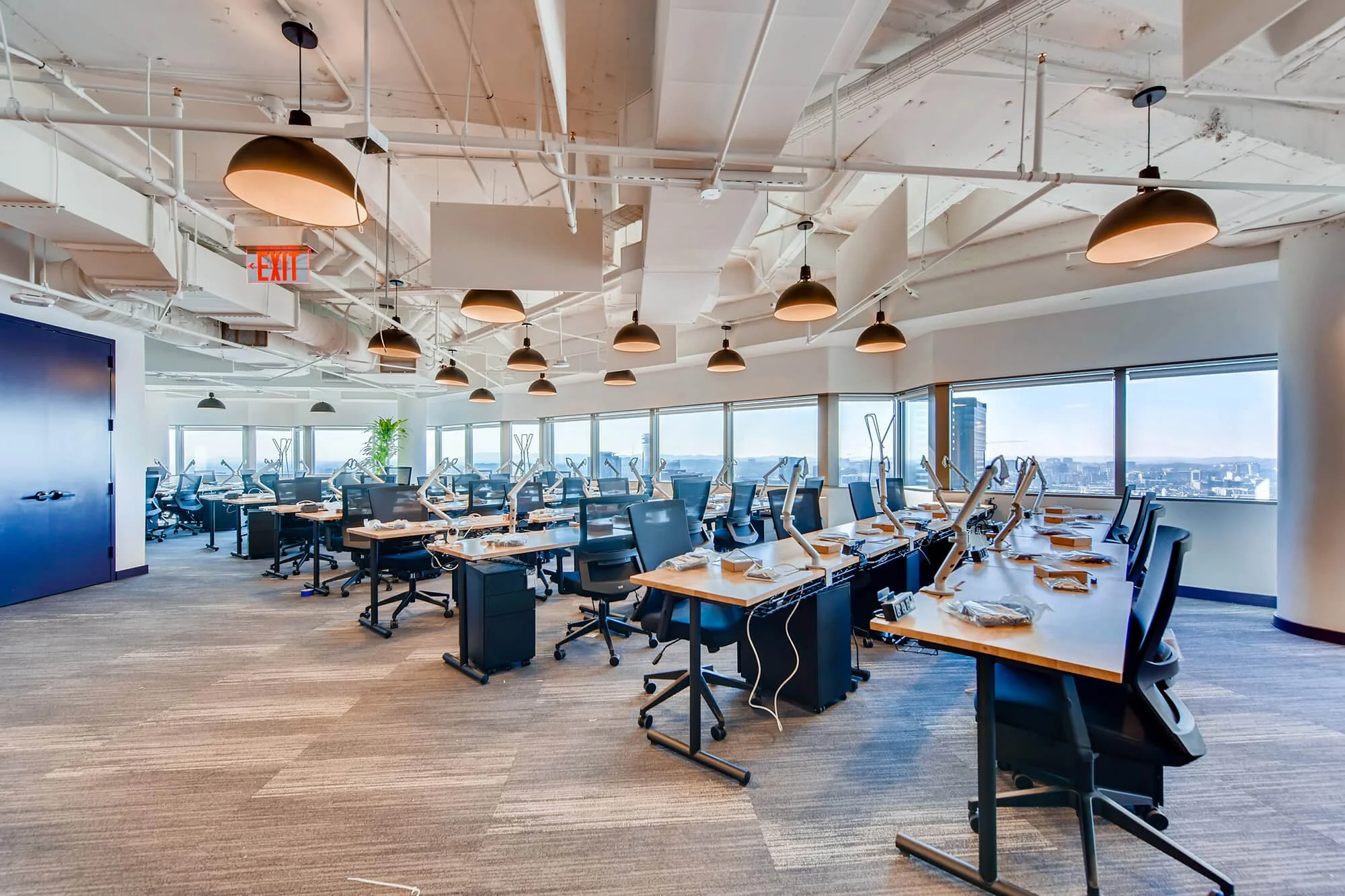  Spacious, modern office interior with rows of desks, chairs, and computer setups, bathed in natural light from large windows overlooking a city skyline, with pendant lights hanging from the exposed ceiling. 