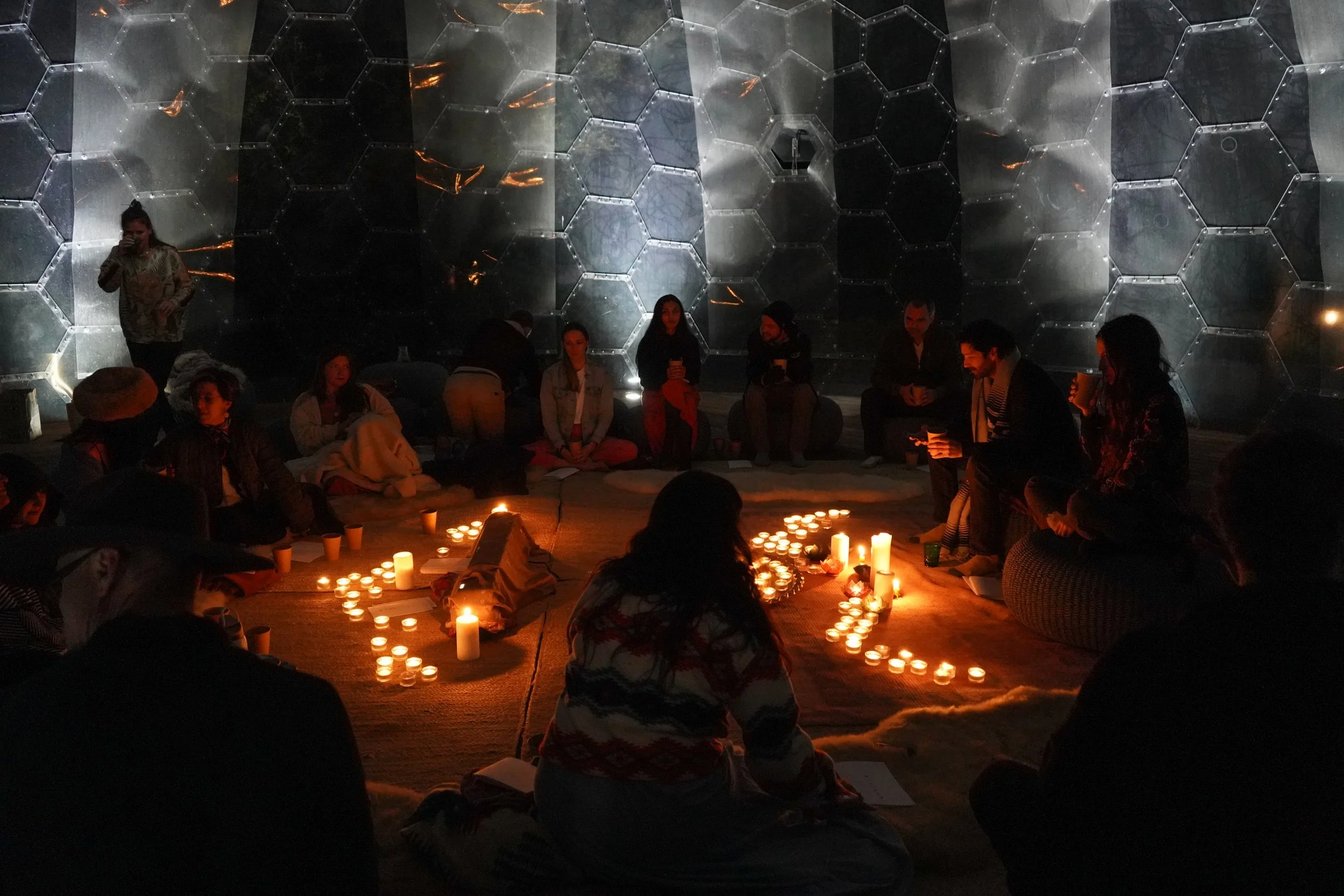 Group of people sitting on the floor around candles, some standing or sitting against a wall, in a dimly lit space with a geometric metal wall background.