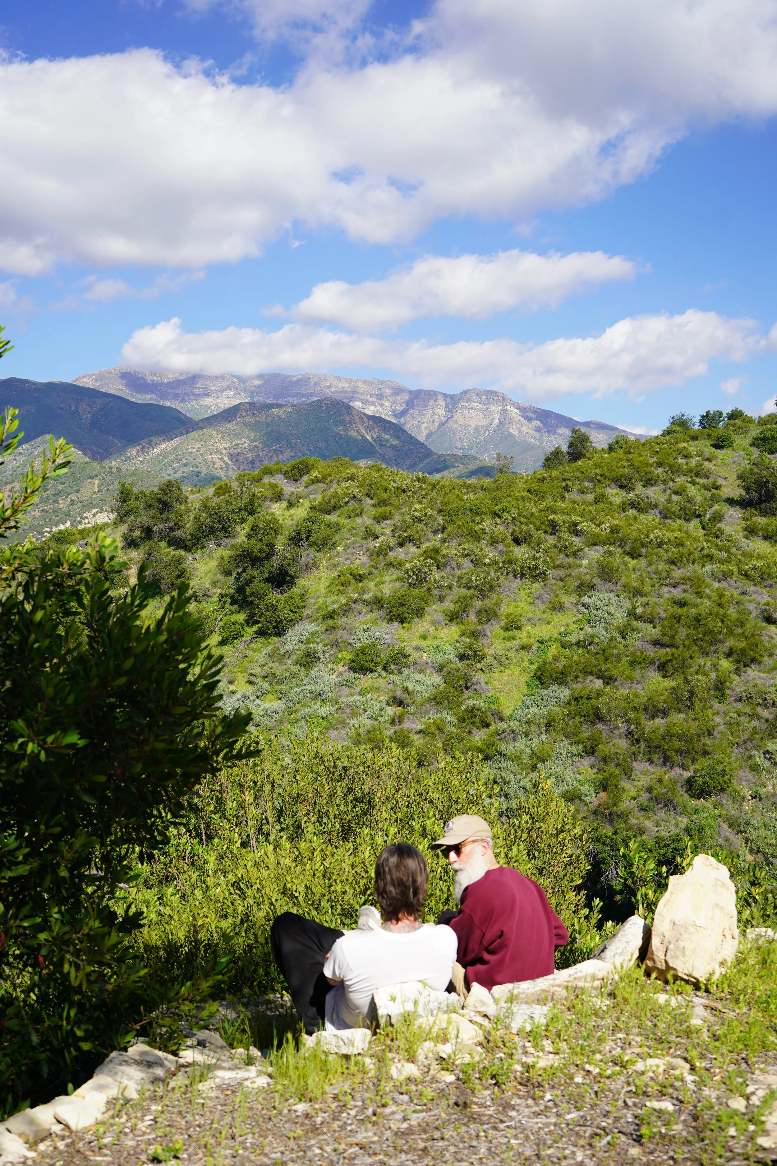 Two people sitting on rocks and conversing in a mountainous outdoor setting with green hills, trees, and blue sky with clouds.