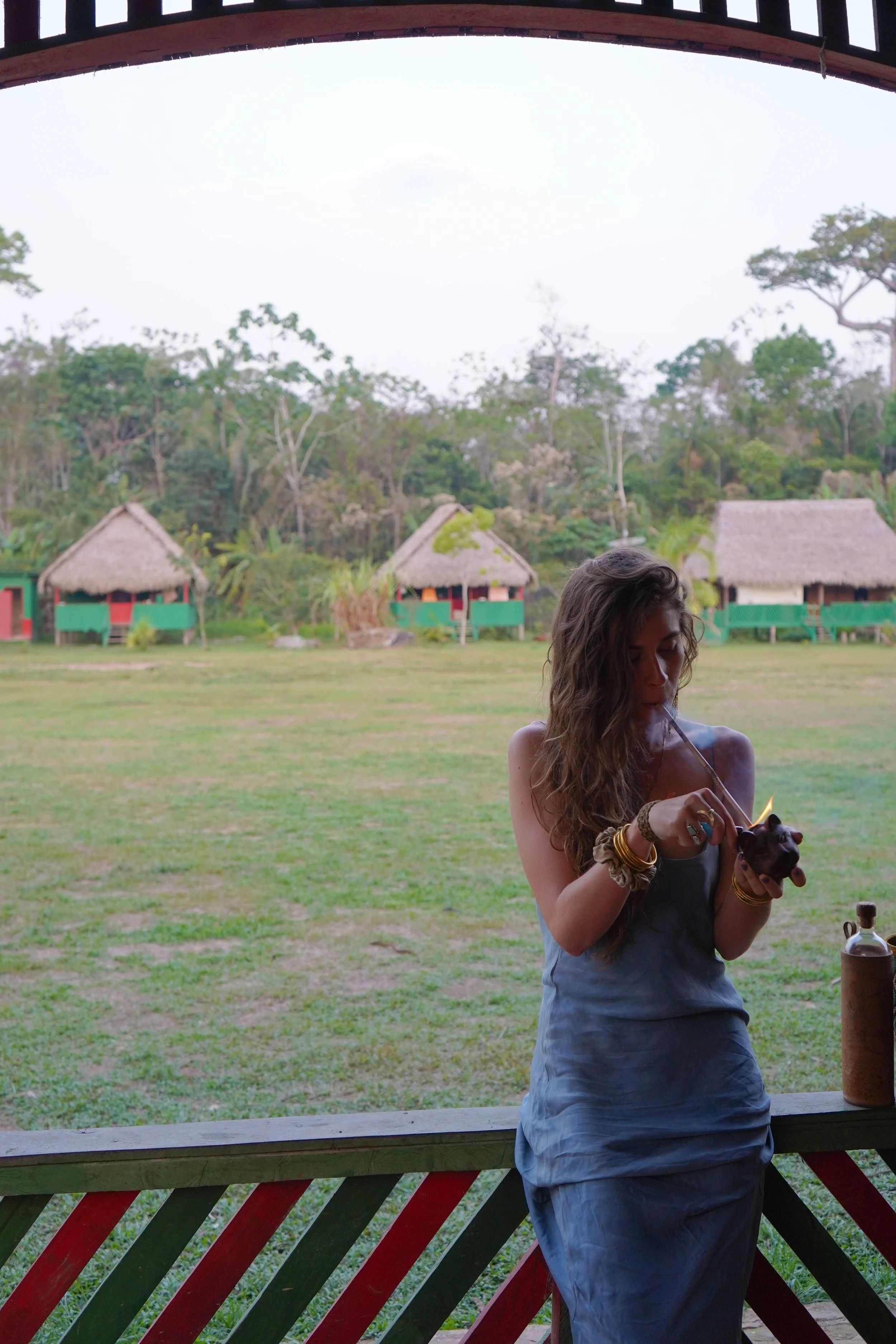 Young woman with long, wavy hair, wearing a gray dress, jewelry, and holding a small object, lighting a pipe with a flame outdoors on a porch, with thatched-roof huts and trees in the background.