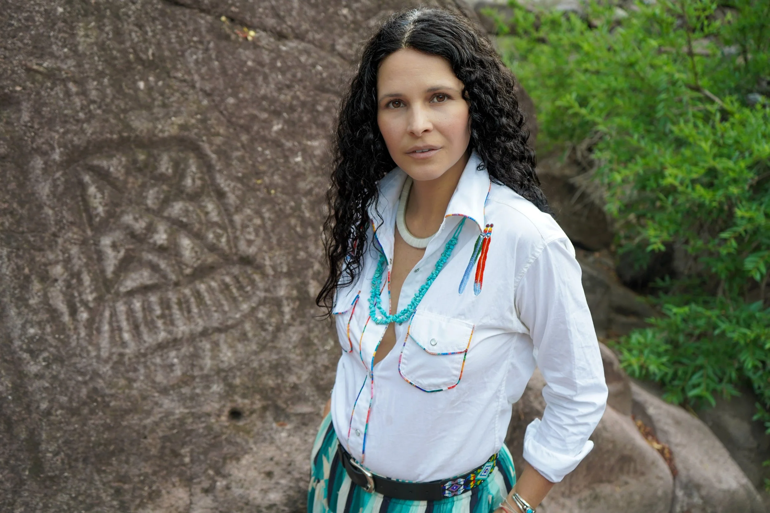 A woman with curly dark hair wearing a white button-up shirt and colorful striped pants stands outdoors in front of a large rock with carvings and green foliage.